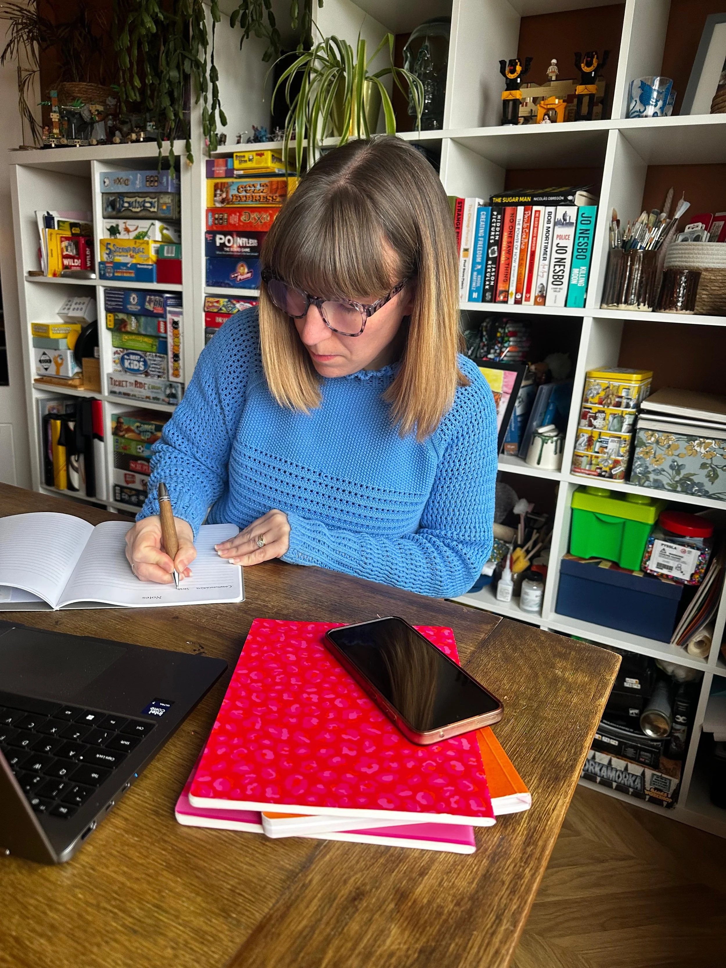 A woman with shoulder-length blonde hair and glasses, wearing a blue sweater, is sitting at a wooden desk writing in a notebook. On the desk are a black laptop, three stacked notebooks with pink, orange, and white covers, and a smartphone. In the background, there is a white bookshelf filled with books, board games, and decorative items.