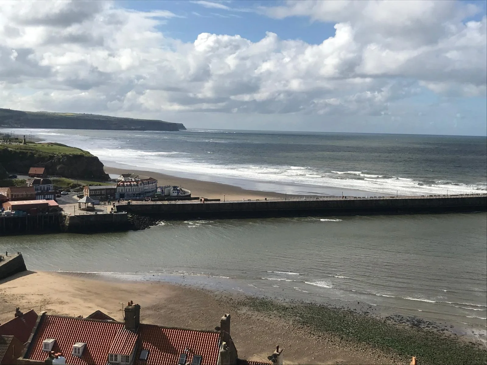 Photograph of the Whitby coastline. Shows a cliff in the background with a pier, beach, sea and rooftops.