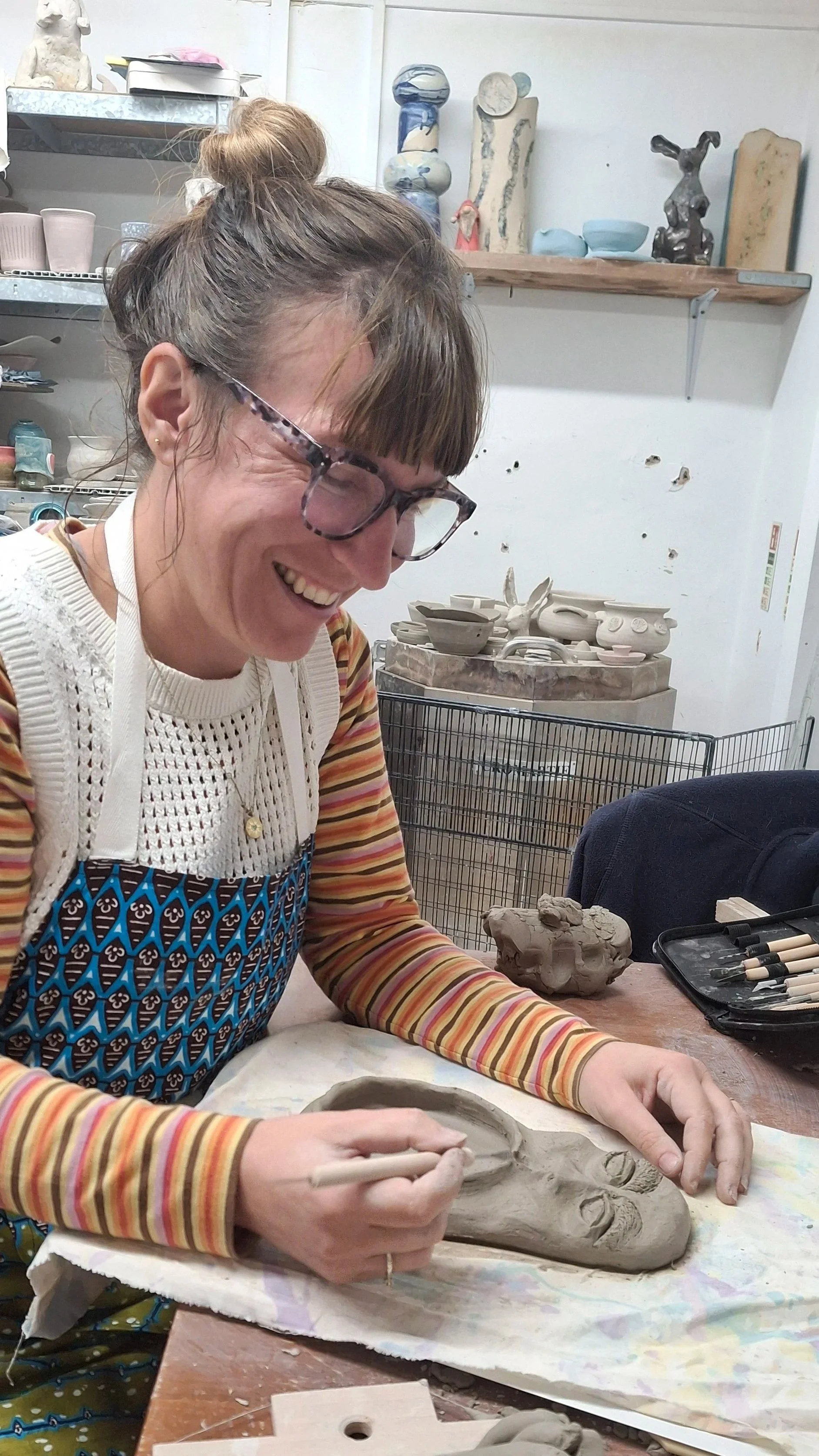 A photo of Kelly wearing glasses and a bun hairstyle joyfully carving a clay sculpture in a ceramics studio.