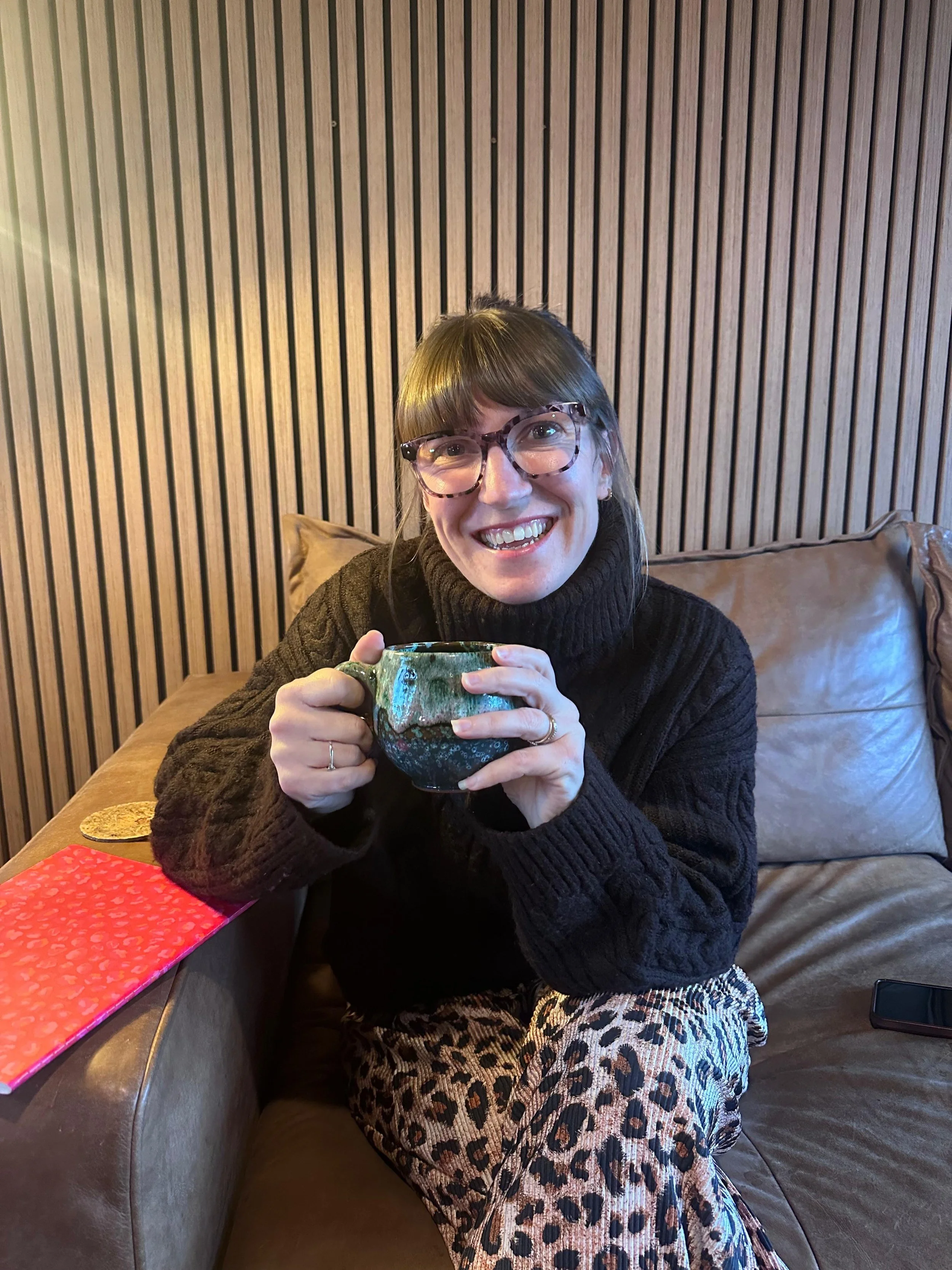 A woman with glasses, wearing a dark sweater and animal print skirt, sitting on a brown leather couch, smiling and holding a green ceramic mug, with a wooden panel wall behind her.