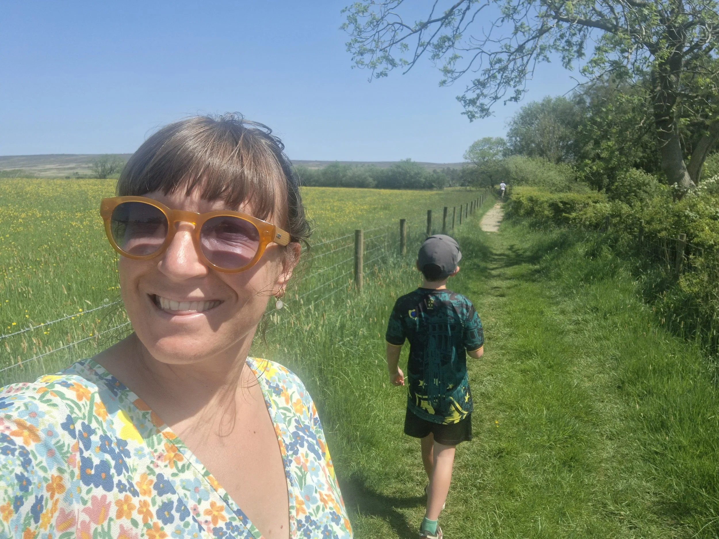 A photo of Kelly smiling at camera wearing sunglasses in the countryside surrounded by fields with child walking away from camera in background.
