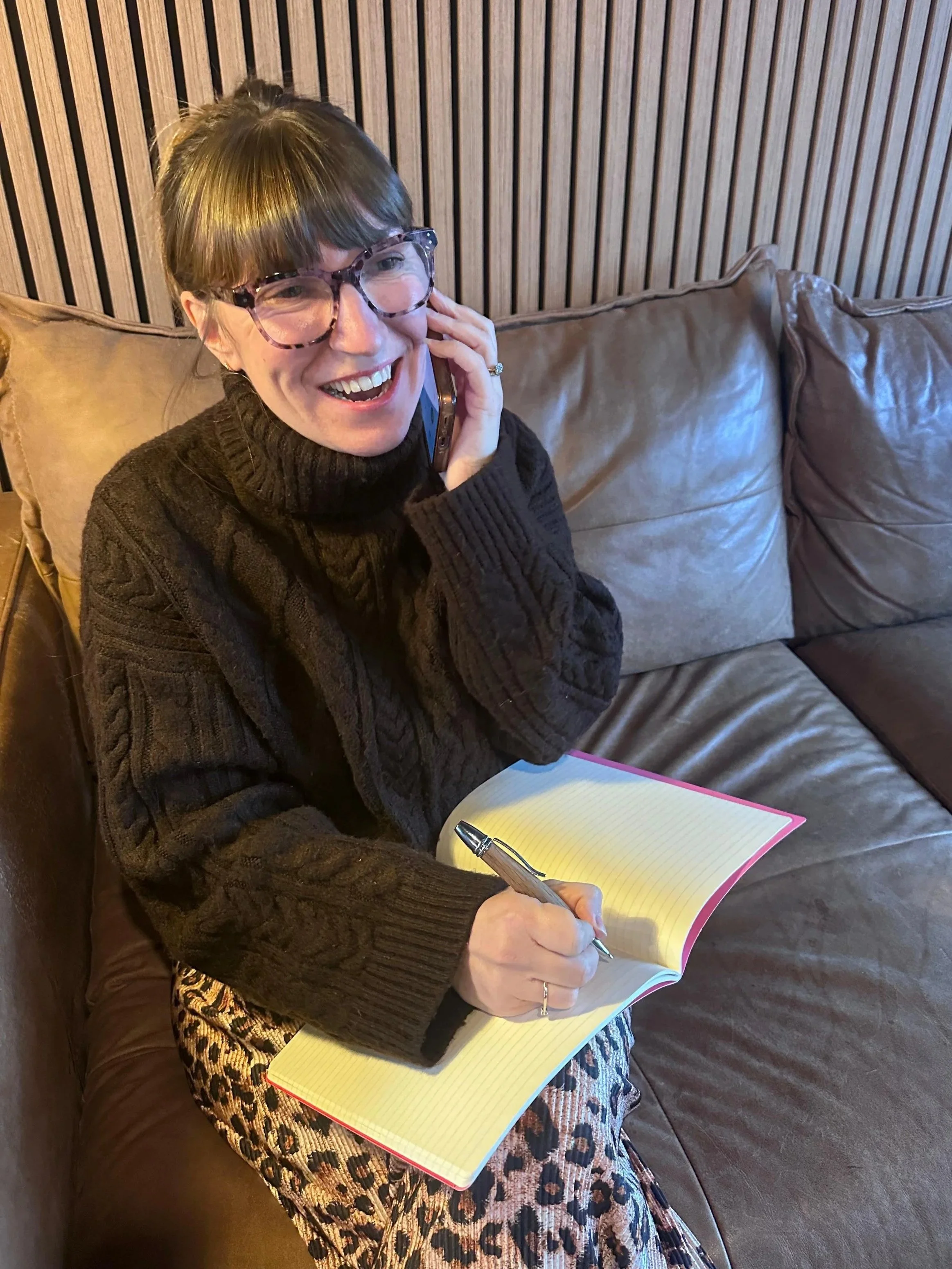 A woman with glasses and brown hair styled in a bun, wearing a dark brown turtleneck sweater, is sitting on a brown leather couch. She is smiling and talking on her cell phone, holding a silver pen and a yellow notepad with a pink border in her lap, with her left hand resting on her face.