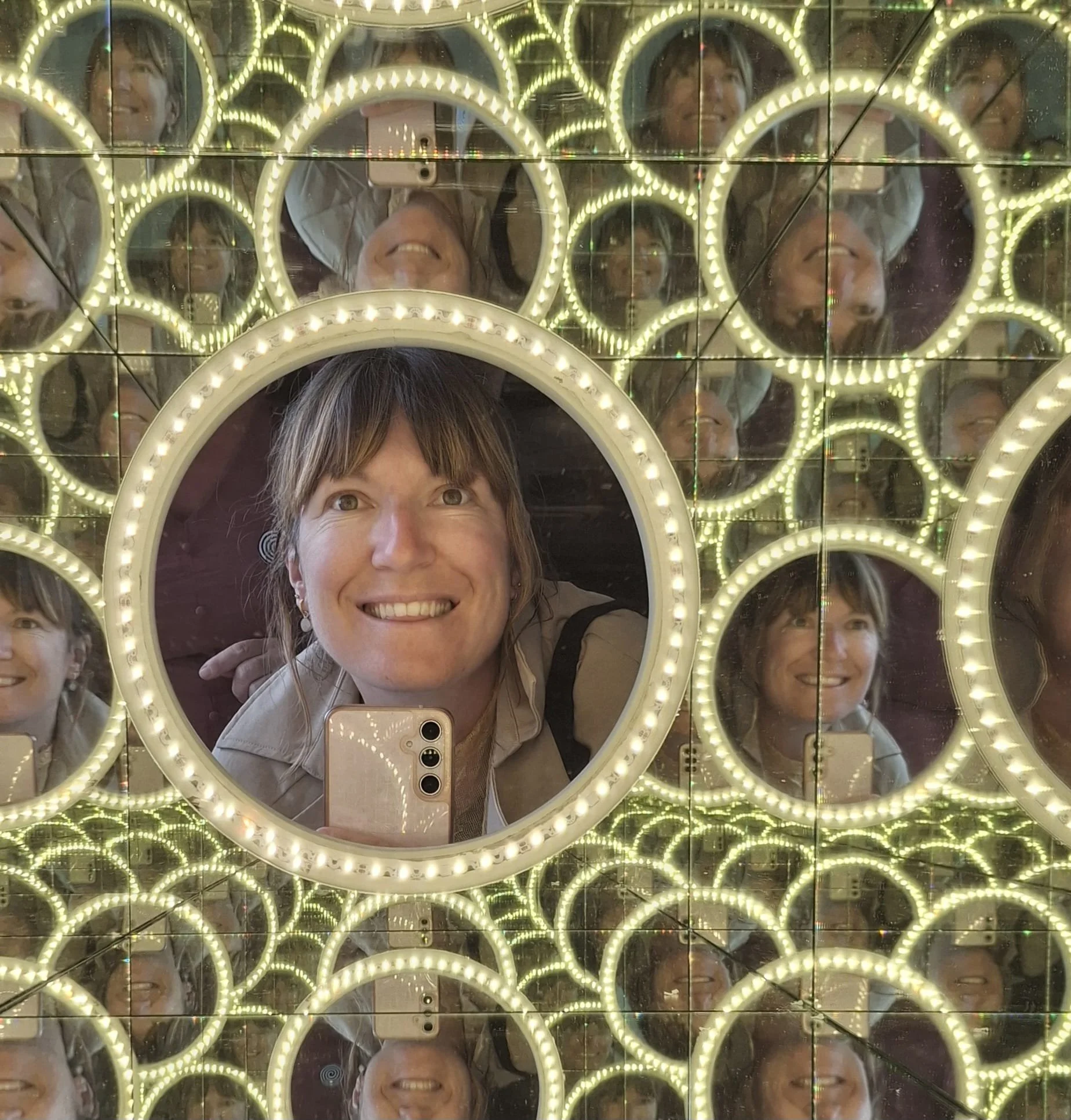 A photo of Kelly taking a selfie in a mirror surrounded by multiple smaller mirrors and circular LED lights, creating a pattern of reflections.