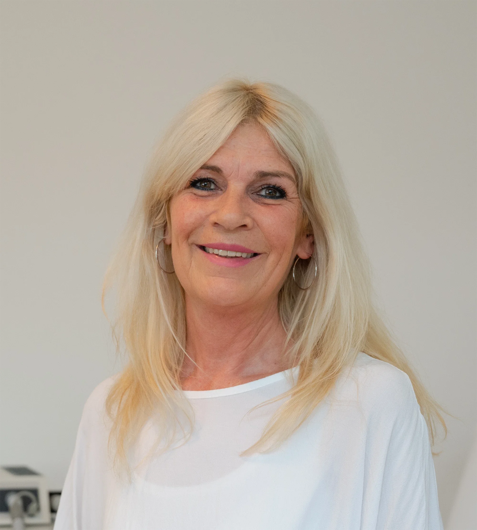 Lächelnde blonde Frau trägt weiße Oberteil, trägt Ohrringe, vor neutralem Hintergrund.