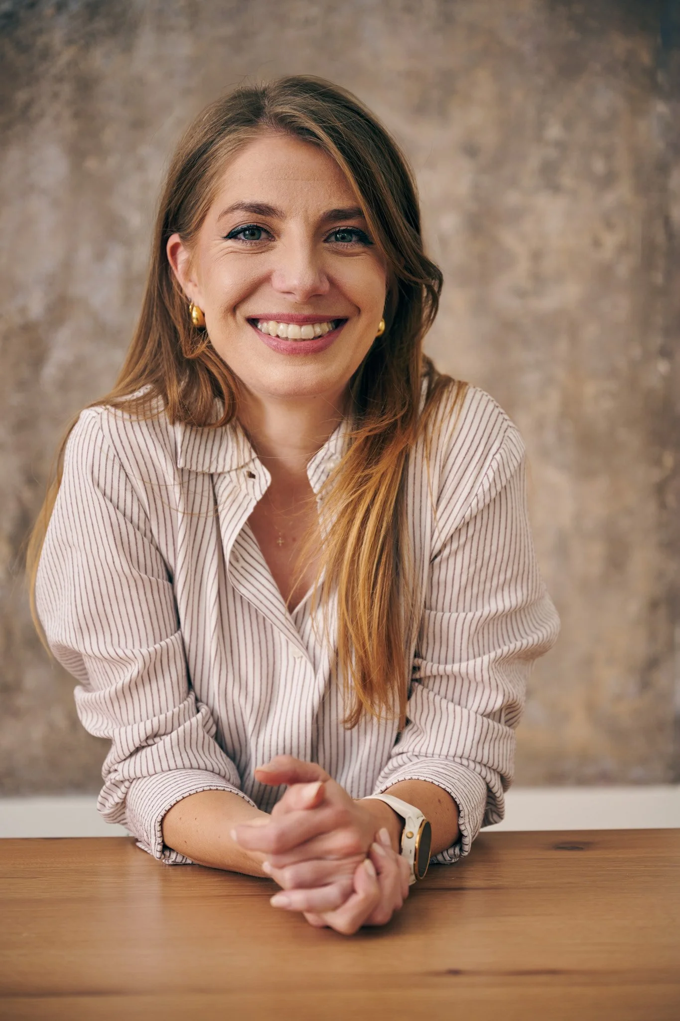 A successful smiling business woman, founder of a business, helping tech businesses grow as a fractional executive, sitting at a wooden table with a neutral, textured background.