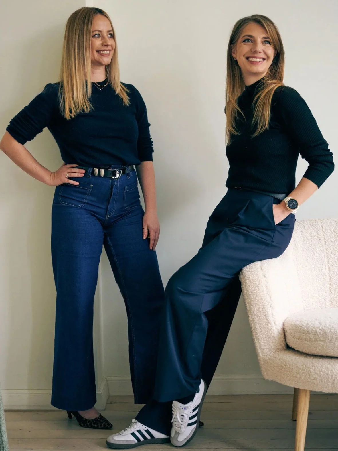 Two women with light skin and long hair smiling and posing in a room with plain white walls. One woman is standing with her hand on her hip, wearing high-waisted jeans, a black top, leopard print heels, and a bracelet. The other woman is sitting on a beige, textured chair, wearing dark pants, a black long-sleeve top, and white Adidas sneakers, with a smartwatch on her left wrist