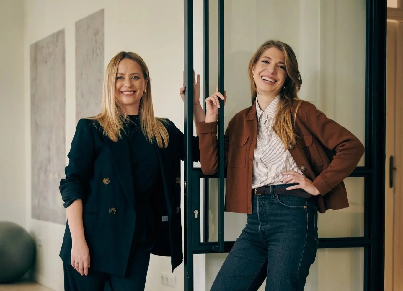Two founders, business women, smiling and posing inside a modern office, modern artwork on the wall, one wearing a dark blazer and the other a brown jacket, standing near a black-framed glass door.
