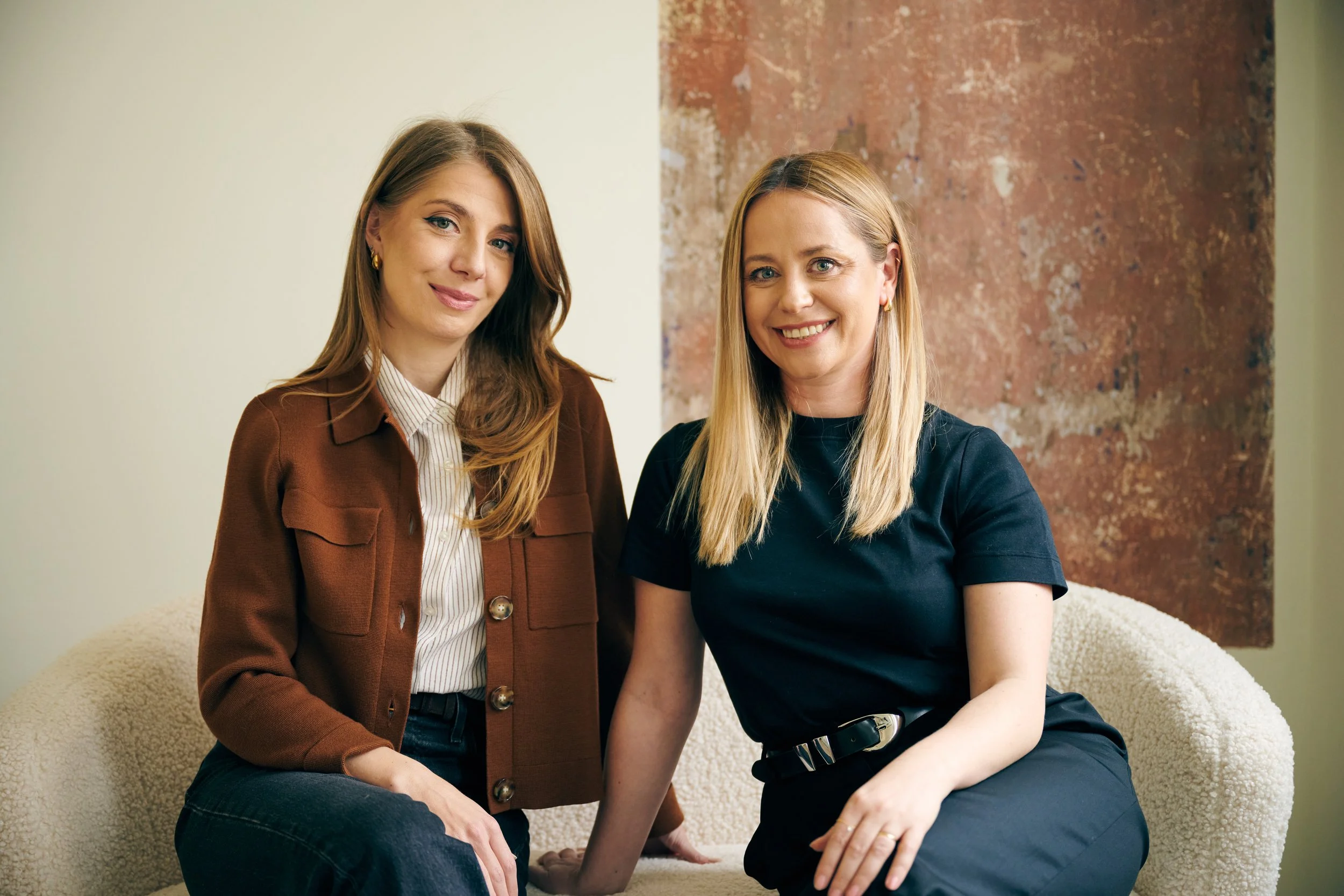 Two women seated on a cream-colored couch, smiling at the camera. The woman on the left has long, light brown hair and is wearing a brown blazer over a striped shirt. The woman on the right has long blond hair and is wearing a black shirt with a black belt.
