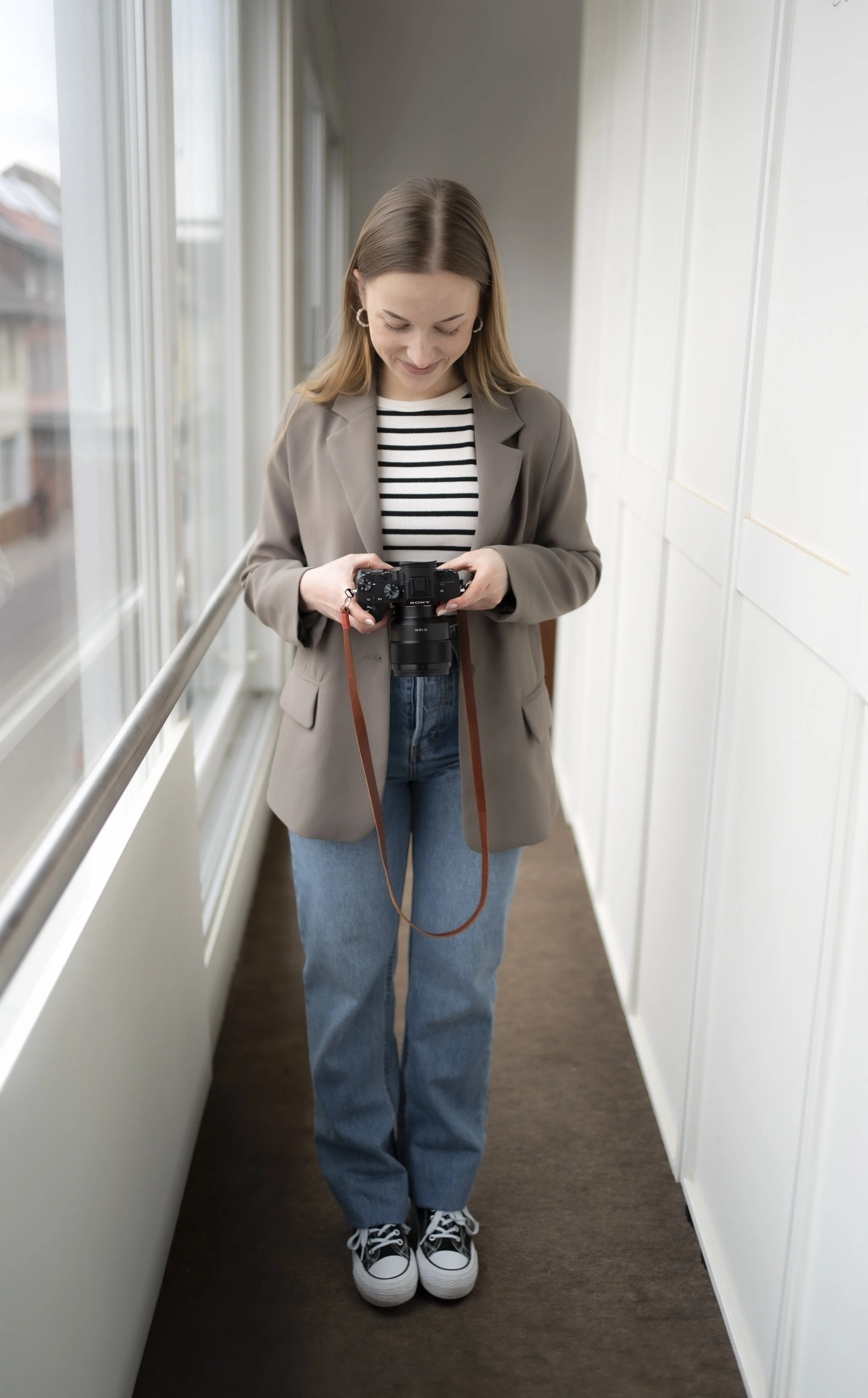 Rebecca Fotografie im grünen Blazer, gestreiftem Shirt, Jeans und Turnschuhen hält eine Kamera in einem hellen Flur mit großen Fenstern und weißen Wänden.