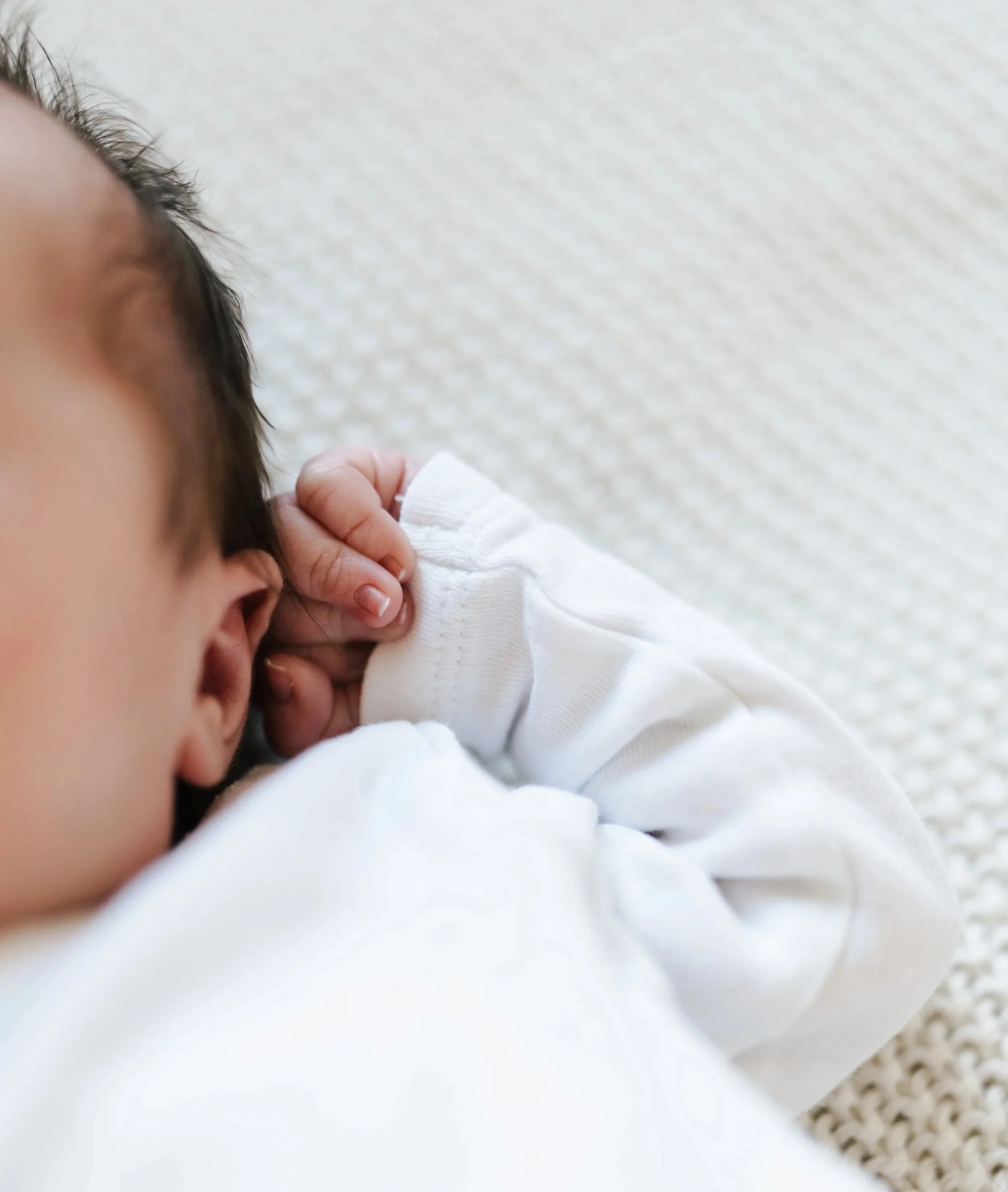 Nahaufnahme eines Babys, das auf einer weißen, strukturierten Oberfläche liegt, mit Fokus auf den Kopf und die Hand, die die Hand zum Ohr hält, während es entspannt liegt. Fotografiert von Rebecca Fotografie in Mühlacker.