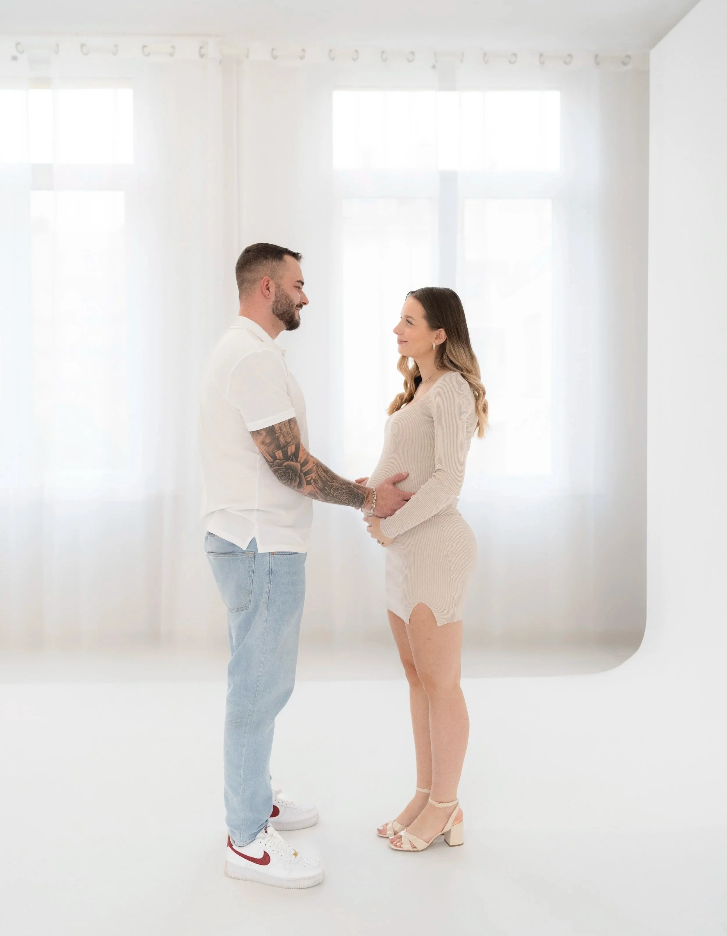 Ein Paar steht sich in einem hellen, weißen Raum gegenüber, die Frau ist schwanger und beide halten ihre Bäuche. Sie schauen sich liebevoll an. Fotografiert von Rebecca Fotografie in Pforzheim in einem Studio.
