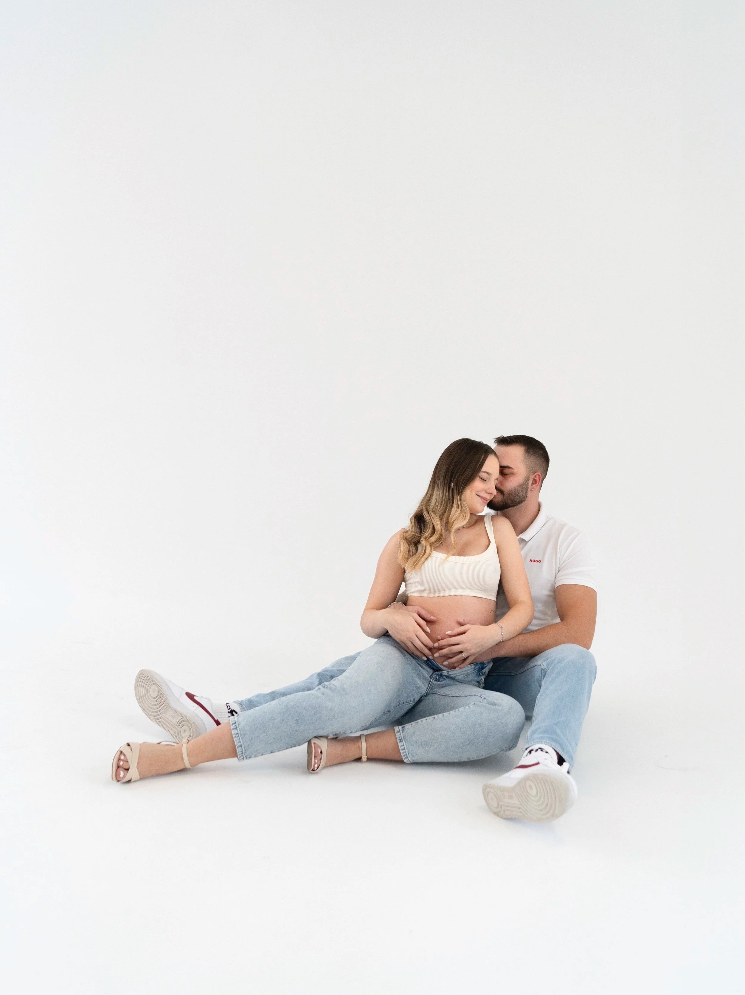 Ein Paar sitzt auf dem Boden, die schwangere Frau ist umarmt vom Mann, beide lächeln liebevoll, weißer Hintergrund. Fotografiert von Rebecca Fotografie in Pforzheim im Fotostudio.