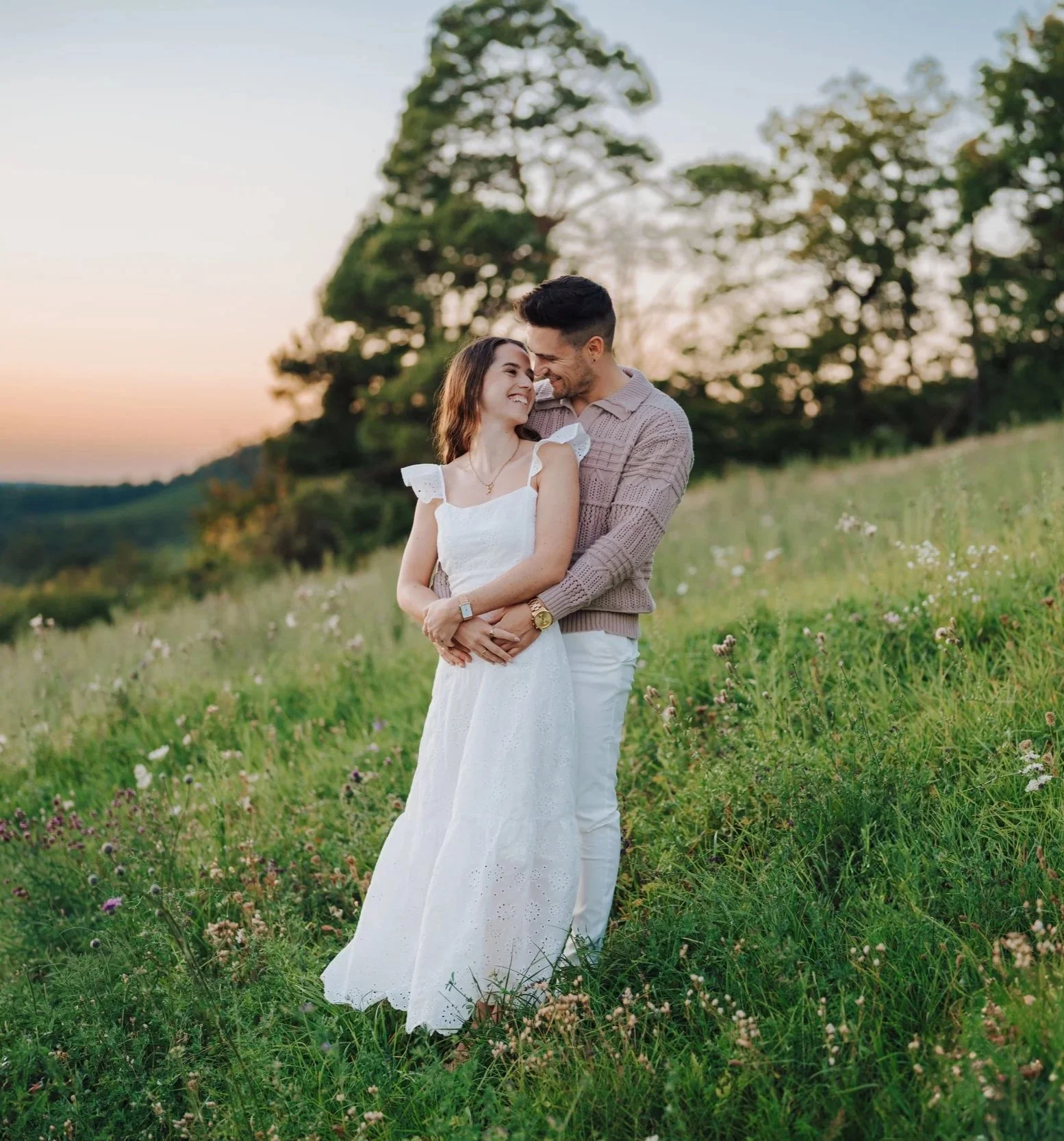 Ein lachendes Paar umarmt sich in einem grünen Feld bei Sonnenuntergang, im Hintergrund Bäume. Fotografiert von Rebecca Fotografie in Vaihingen-Enz.
