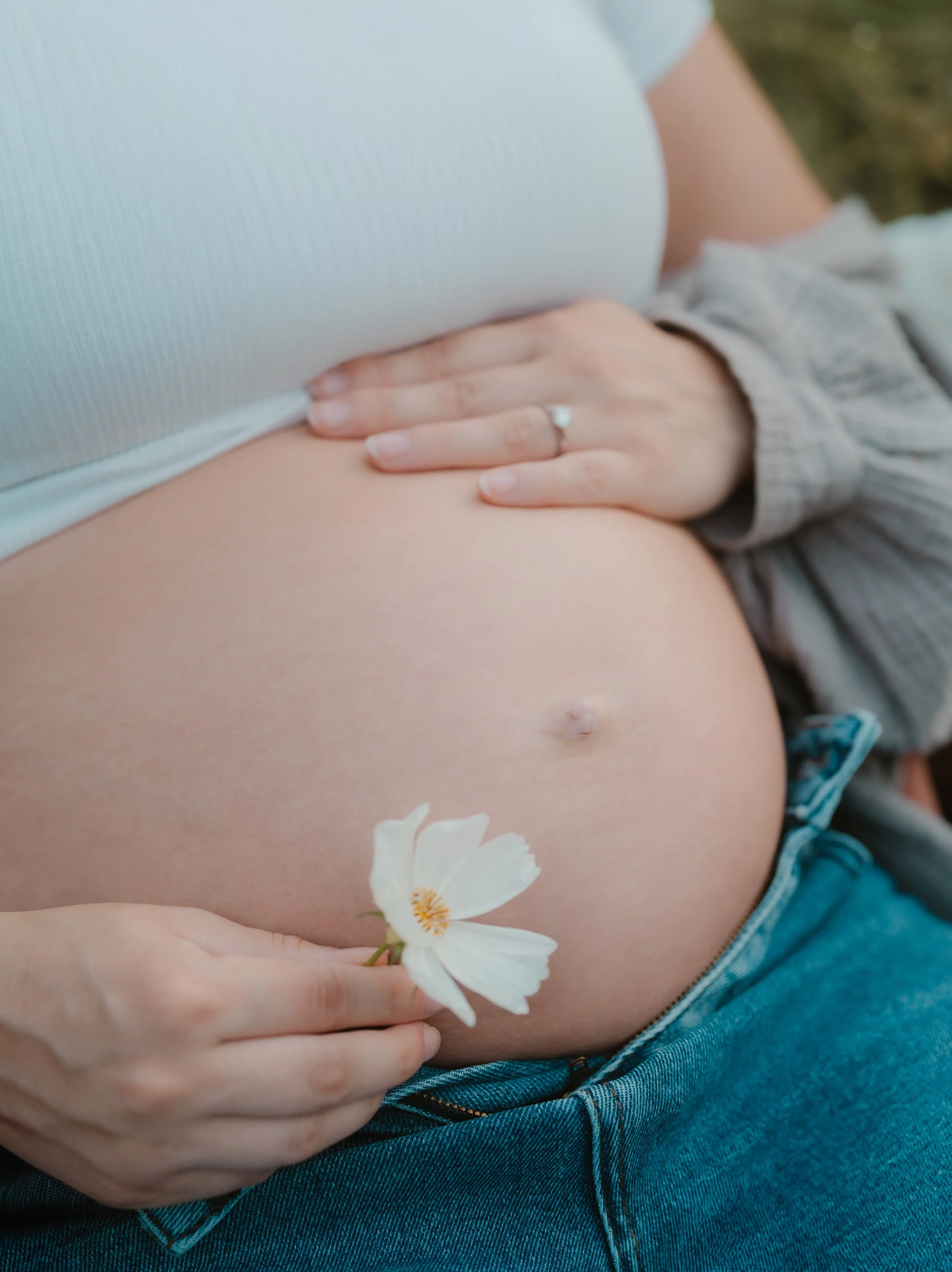 Nahaufnahme eines schwangeren Bauchs, eine Hand hält eine weiße Blume, eine andere Hand liegt auf dem Bauch, im Hintergrund unscharf, draußen. Fotografiert von Rebecca Fotografie in Bretten.