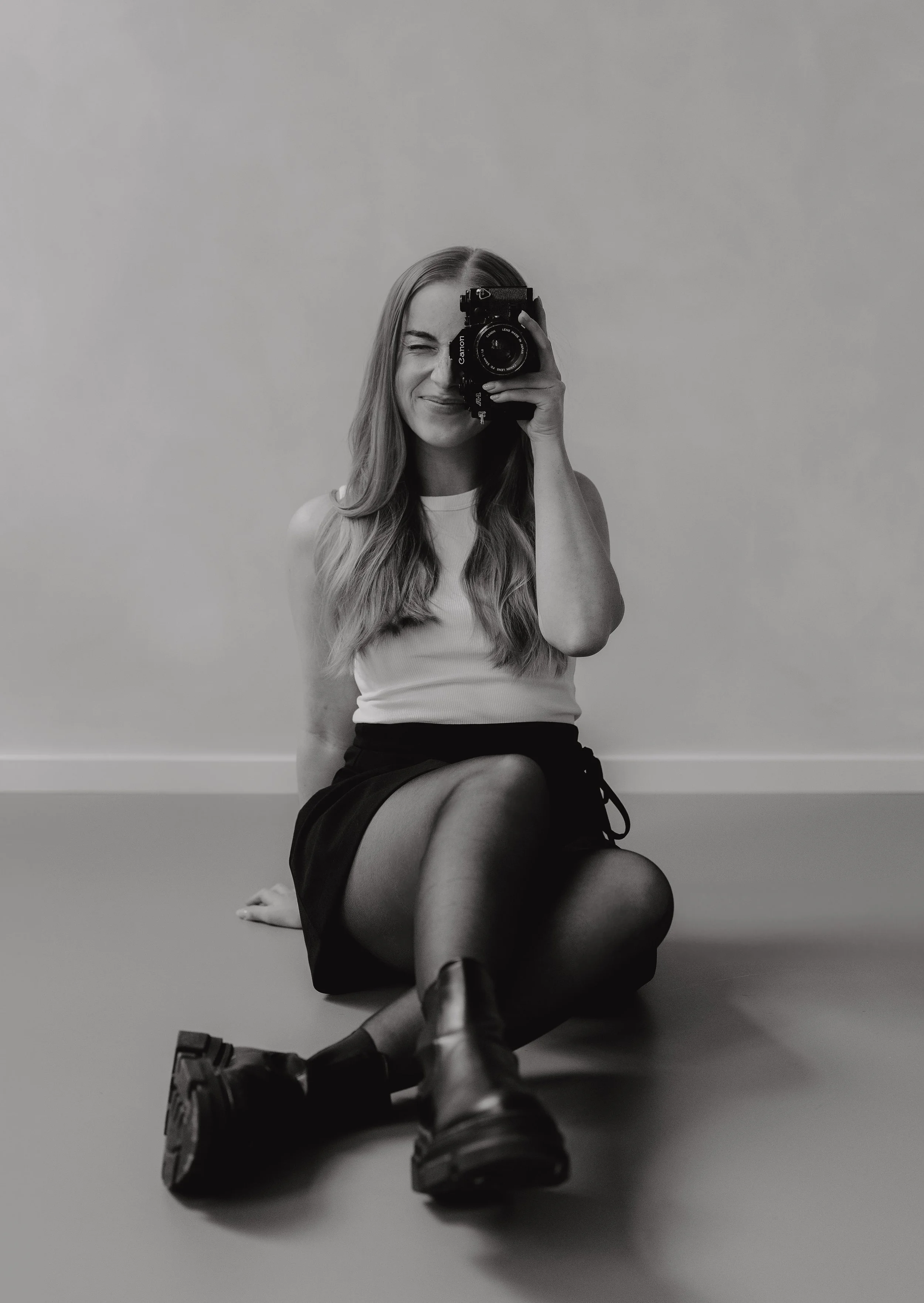 Rebecca  sitzt auf dem Boden, macht ein Selfie mit einer Kamera, trägt ein weißes Top und einen schwarzen Rock, schwarze Strumpfhose und Stiefel, im Schwarz-Weiß-Foto in einem Fotostudio.