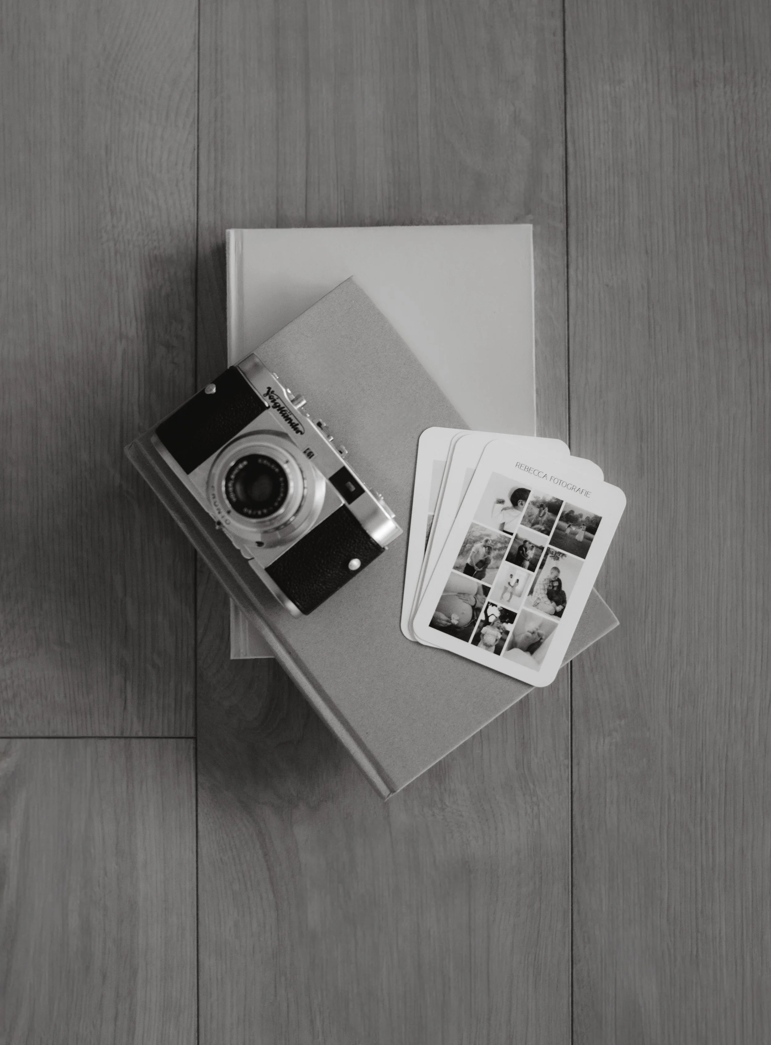 Eine Kamera, ein Fotoalbum und Fotokarten auf einem Holzfußboden und ein Flyer von Rebecca Fotografie.