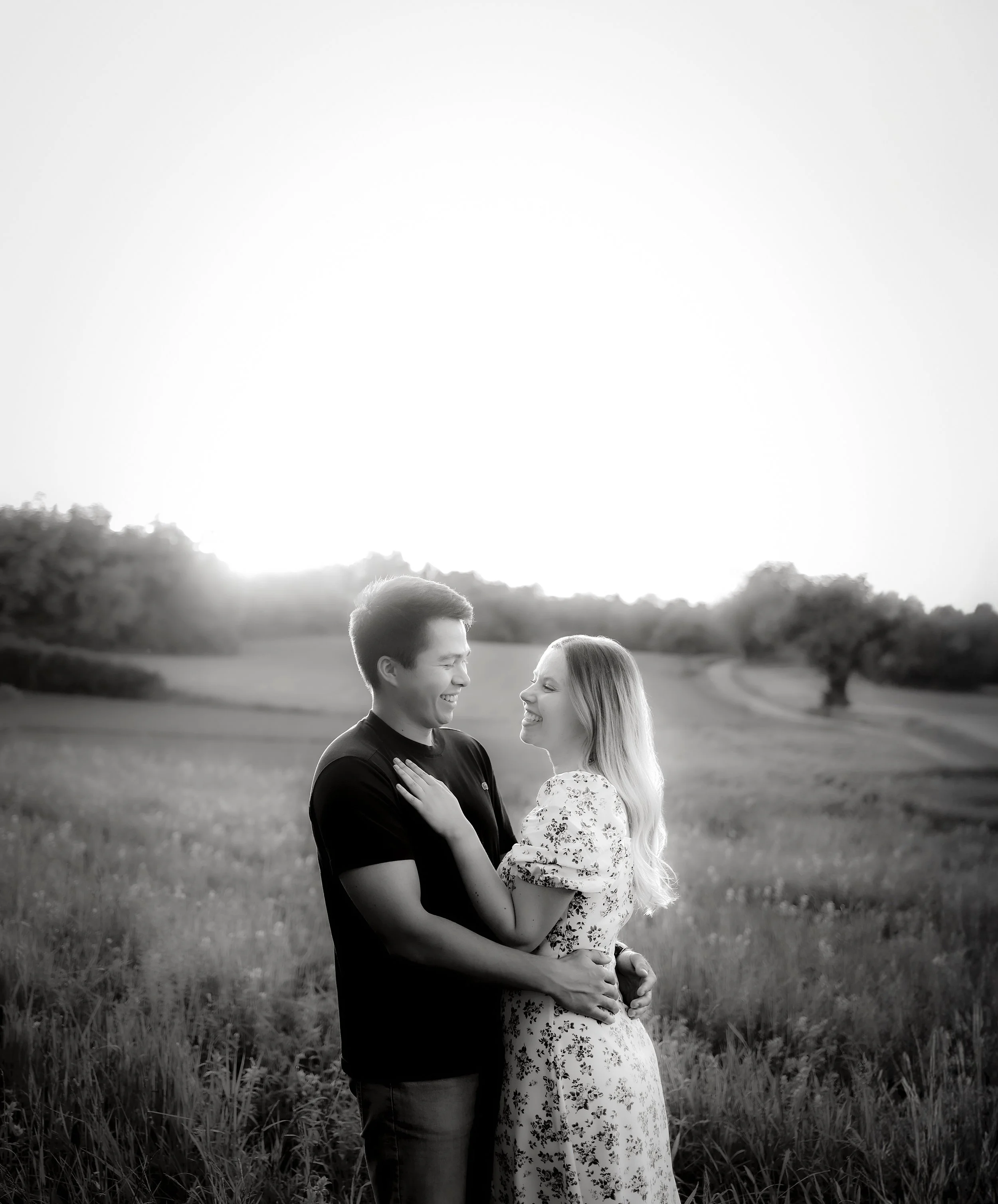 Ein Paar steht in einem Feld bei Sonnenuntergang, lächelt sich an und hält sich umarmt, schwarz-weiß Fotografie. Fotografiert von Rebecca Fotografie in Bretten.