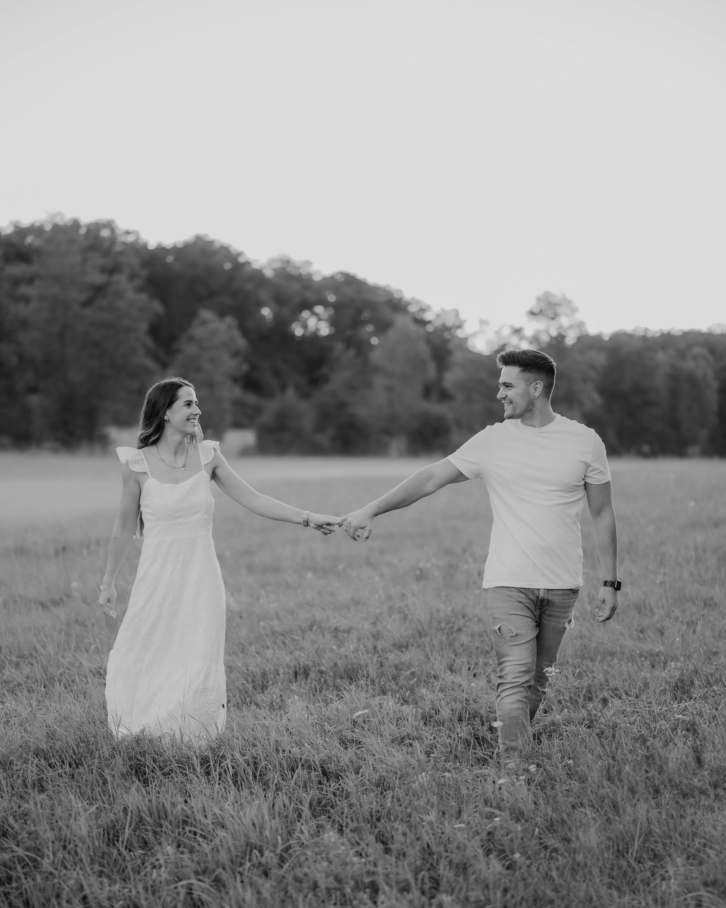 Ein Paar hält sich an einem Feld draußen an einem sonnigen Tag, beide lächelnd und in Richtung einander blickend. Fotografiert von Rebecca Fotografie in Mühlacker.