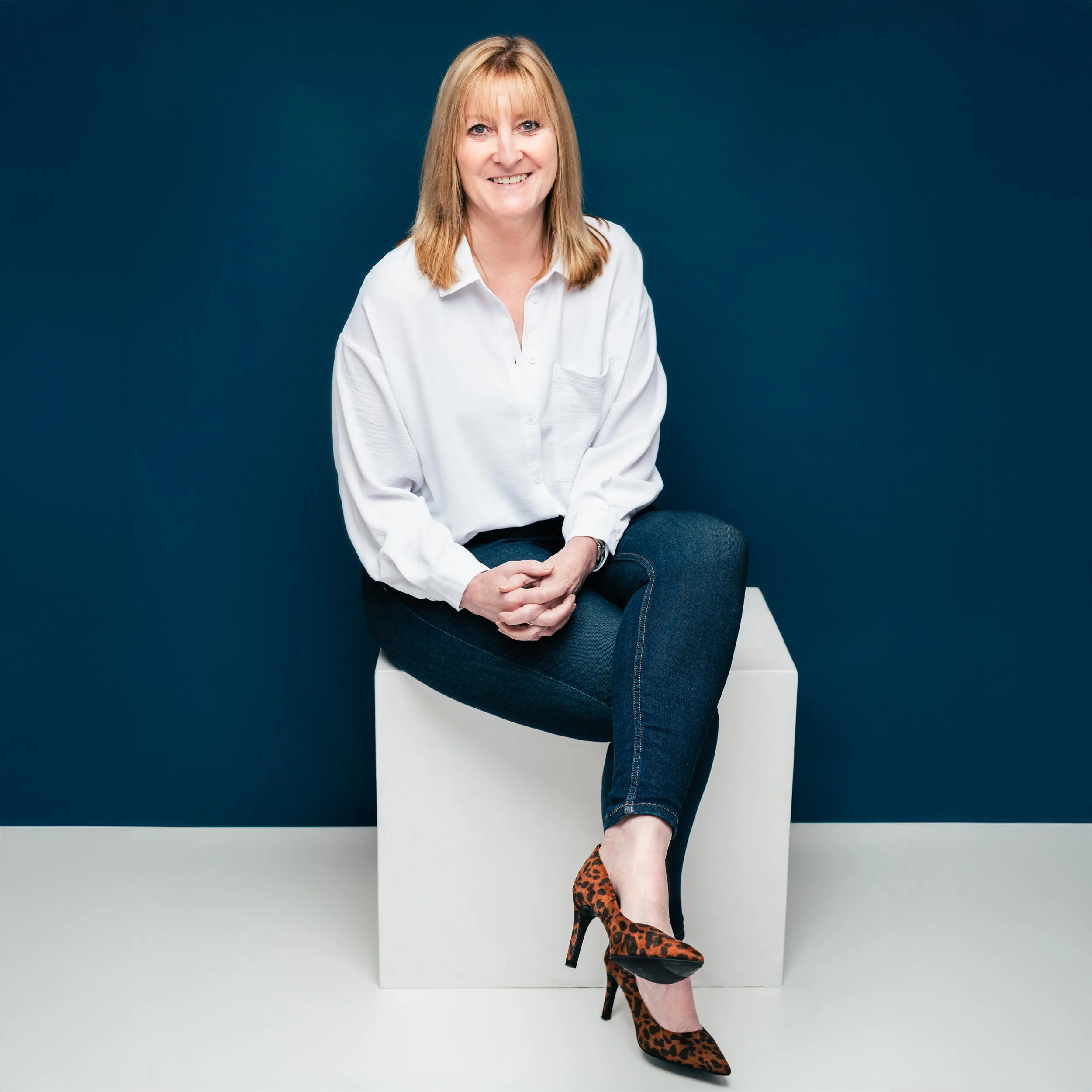 A woman with shoulder-length red hair, wearing a white blouse, dark blue jeans, and leopard print high heels, sitting on a white block against a dark blue background.