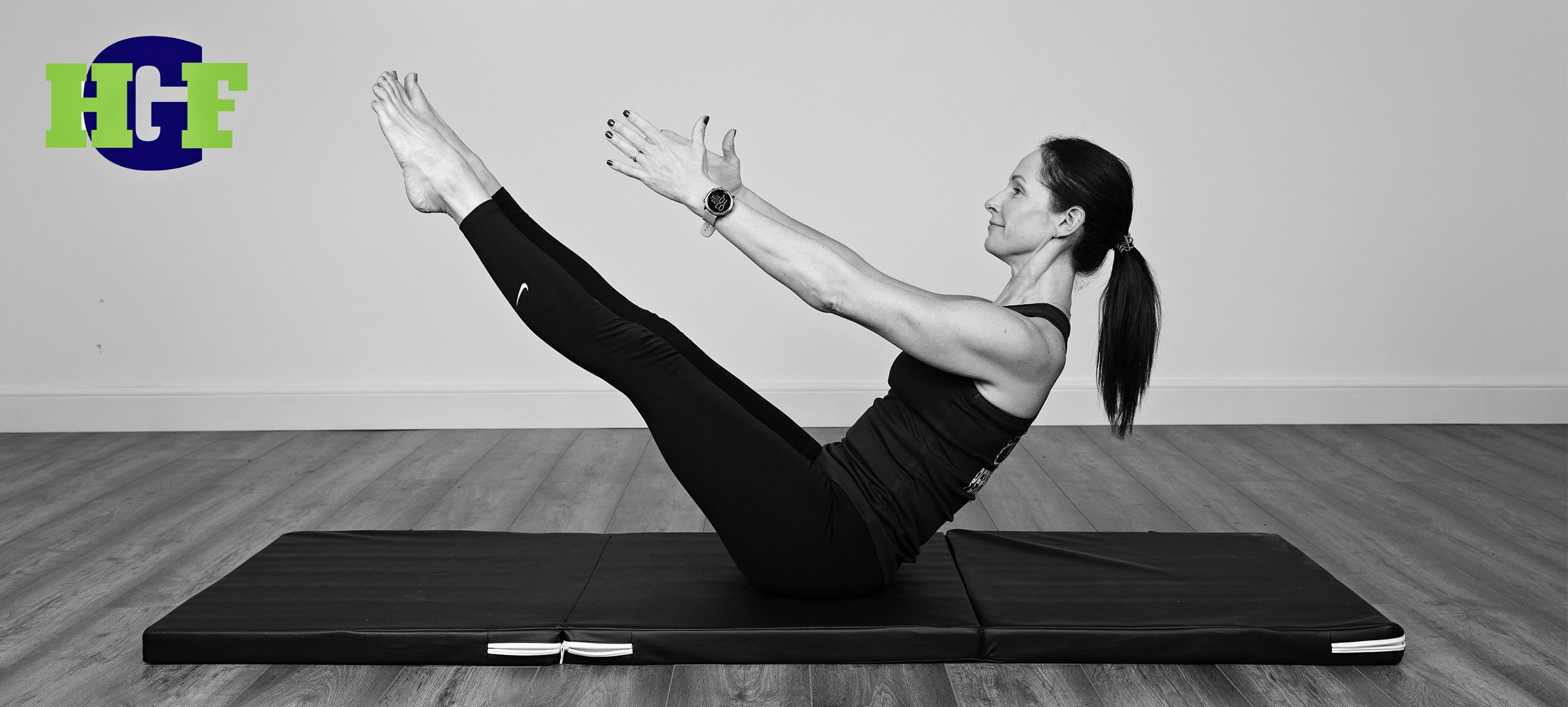 A woman practicing Pilates on a black mat, sitting with legs extended and reaching forward with arms, in a room with wood flooring and plain wall.