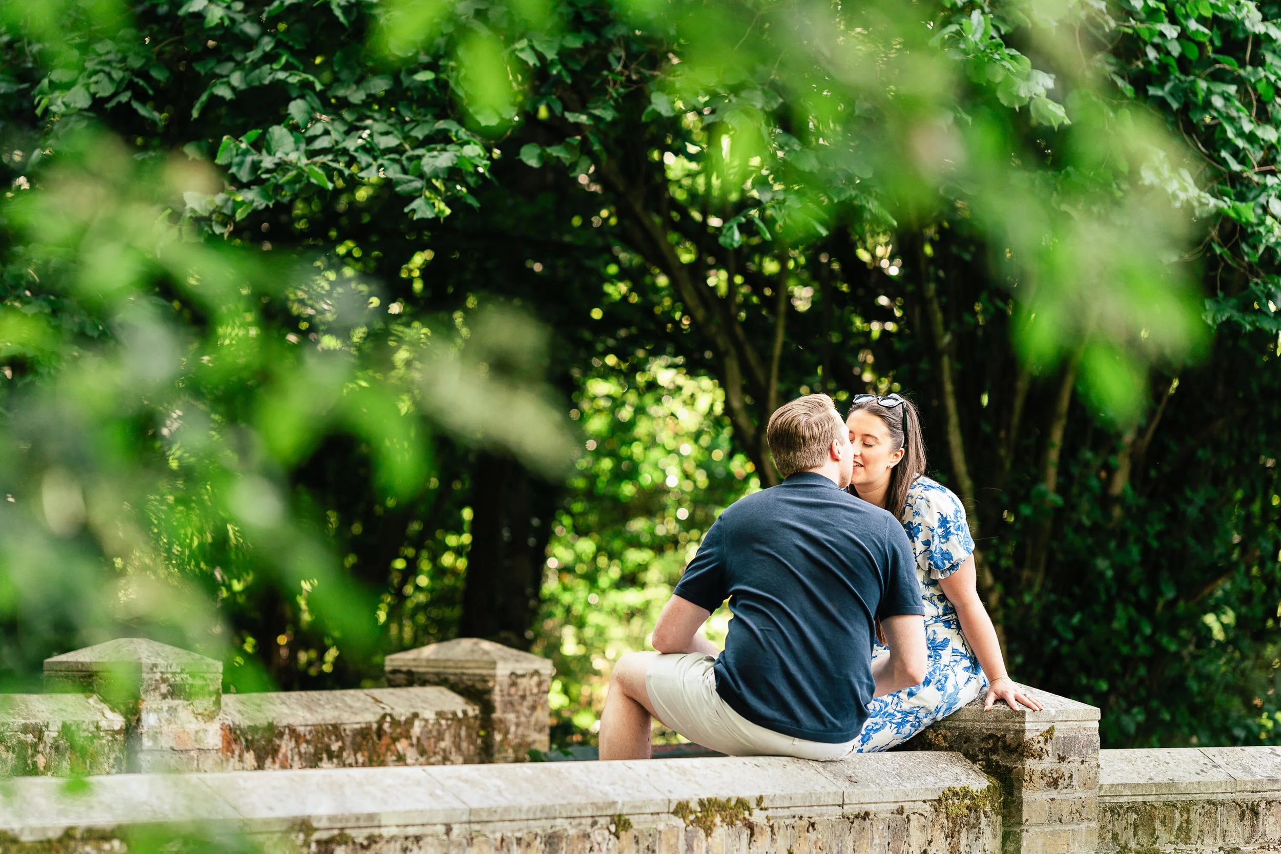 A couple sharing a kiss while sitting on a stone wall in a lush green park, surrounded by trees and foliage.