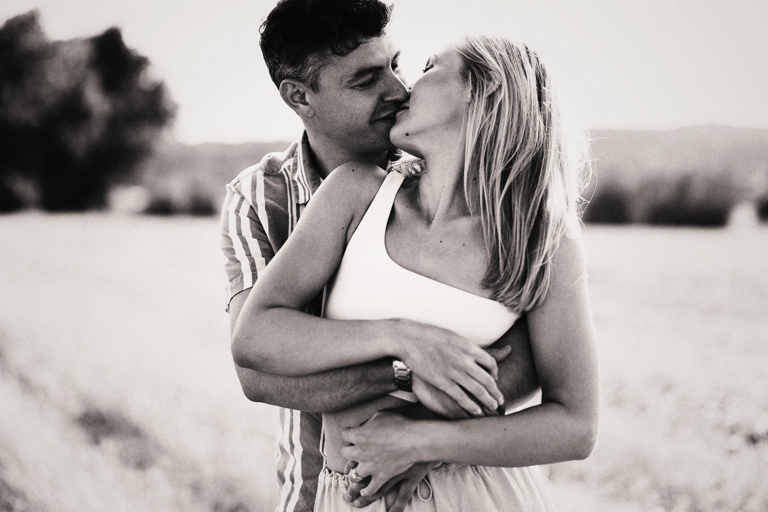 A black and white photo of a couple kissing outdoors, with the man holding the woman from behind.