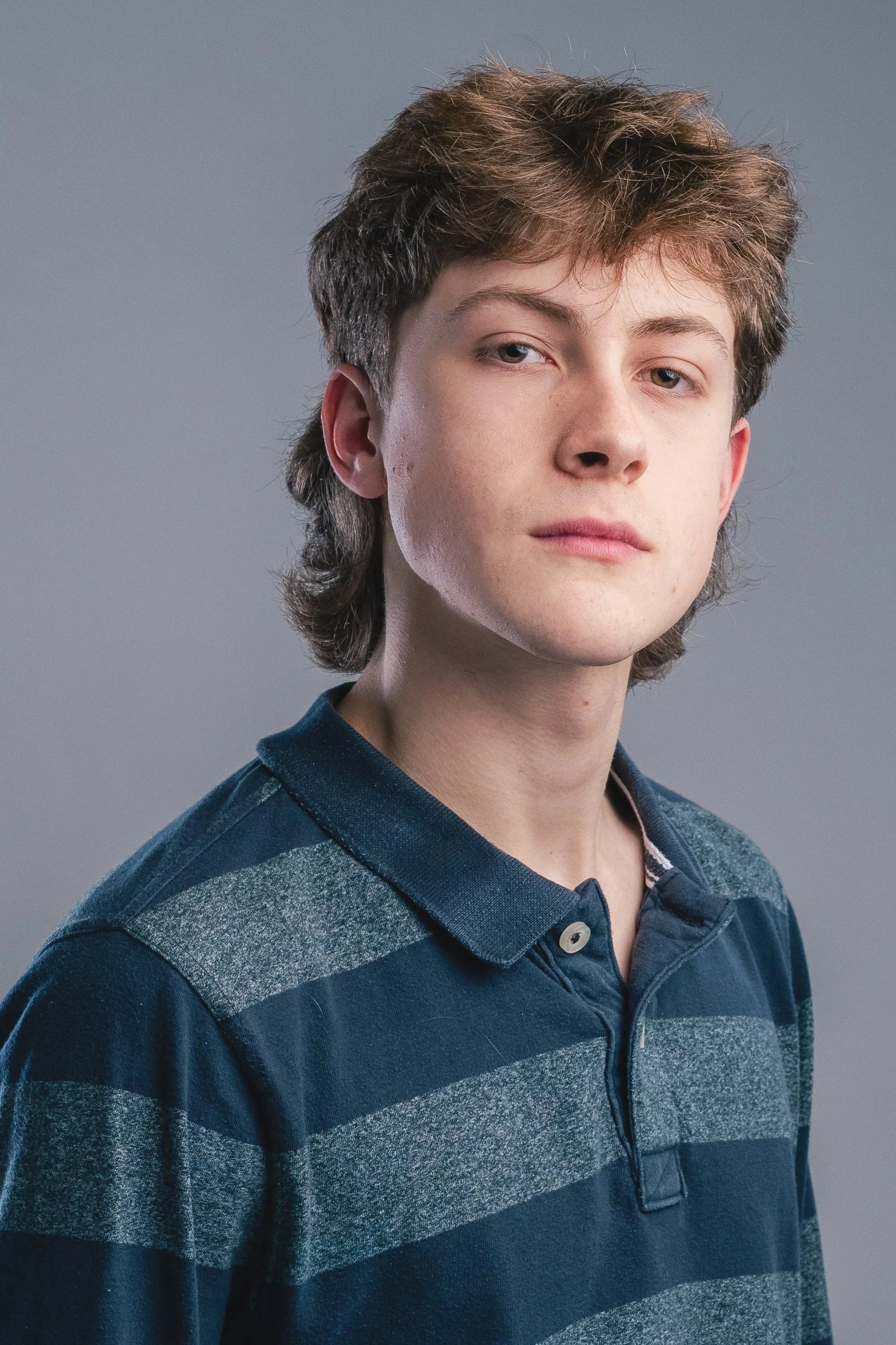 A young man with light skin and brown, tousled hair wearing a blue and gray striped polo shirt, looking slightly serious against a plain gray background.