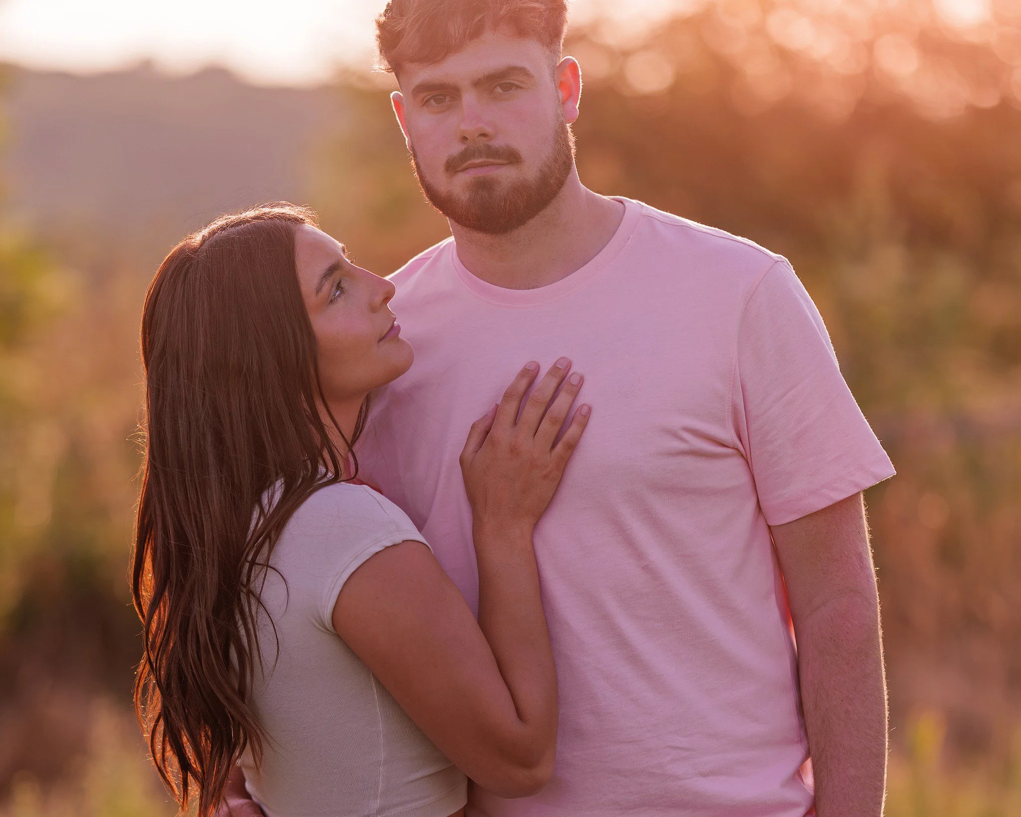 A couple standing outdoors at sunset, with the woman touching the man's chest and looking up admiringly at him.