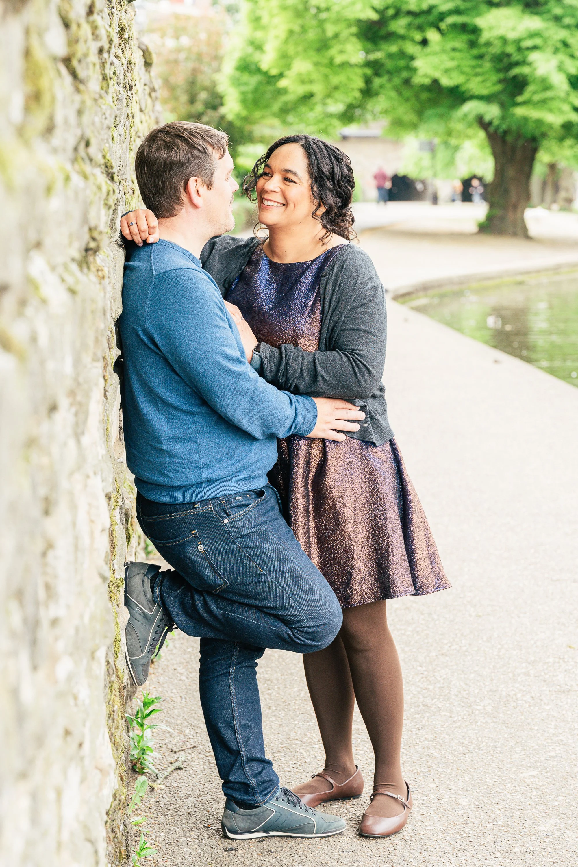 A couple standing by a stone wall in a park, smiling and looking at each other, with trees and a pond in the background.