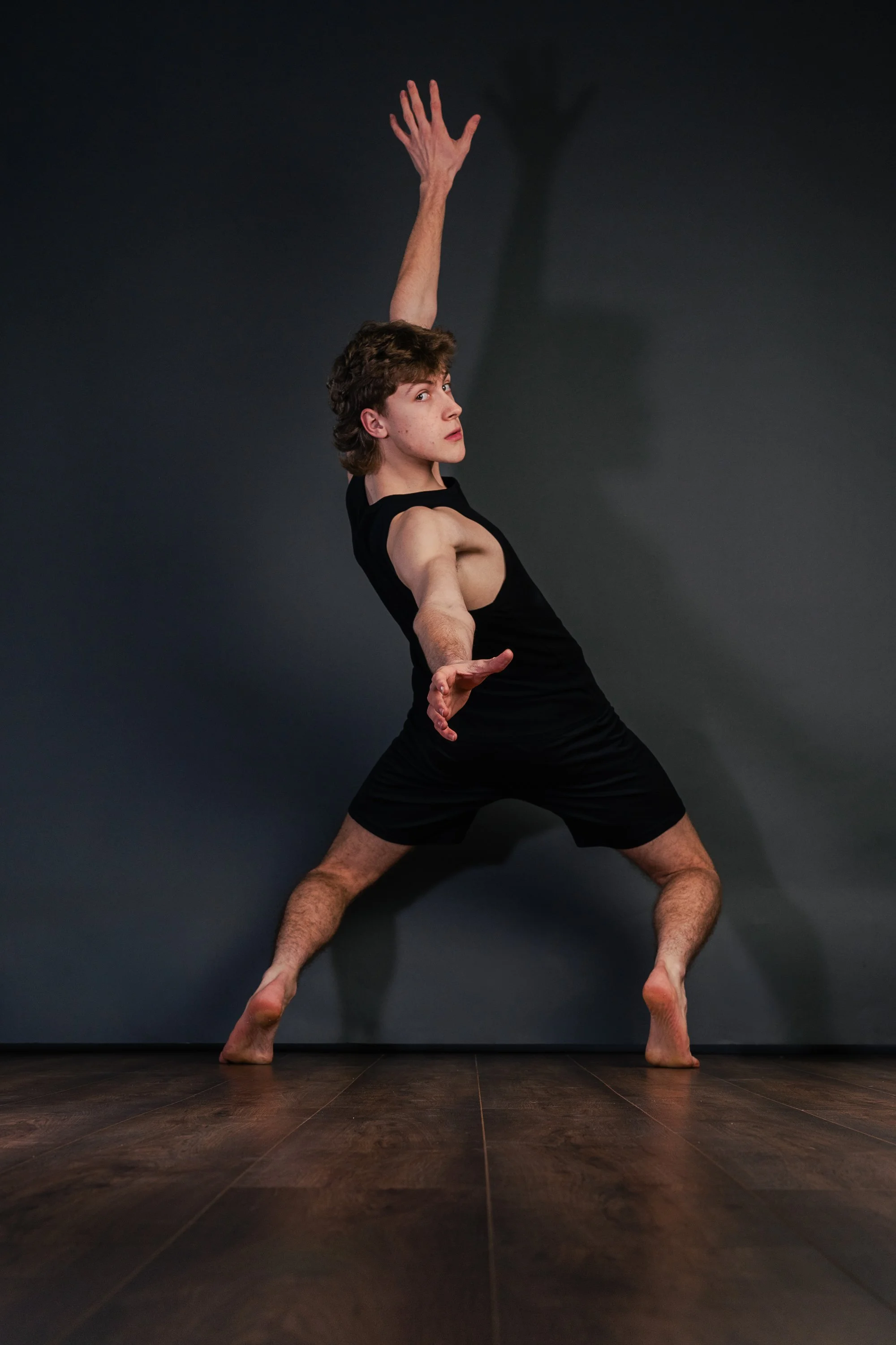 A person in black clothing performing a dance or yoga pose against a dark background, with shadow cast on the wall.