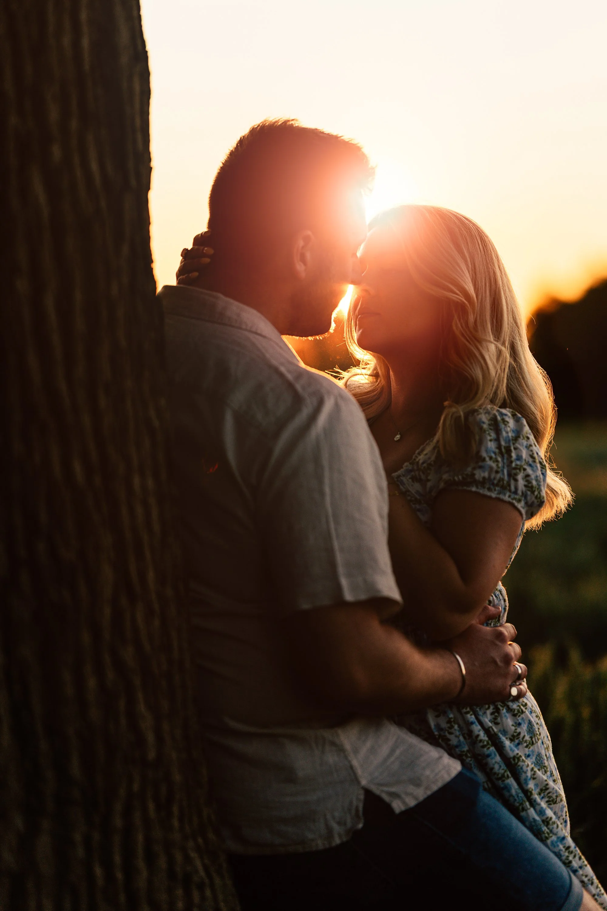 A couple leaning against a tree at sunset, about to kiss, with the sun setting behind them.