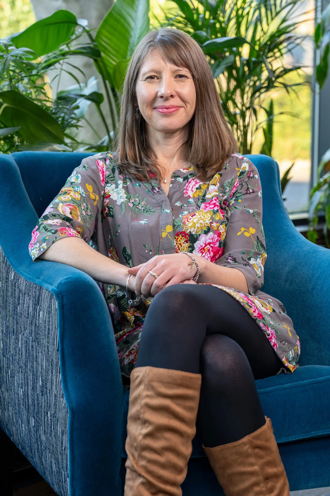 A woman with shoulder-length brown hair, smiling, sitting in a blue armchair in an indoor space with large green leafy plants and a glass window in the background.