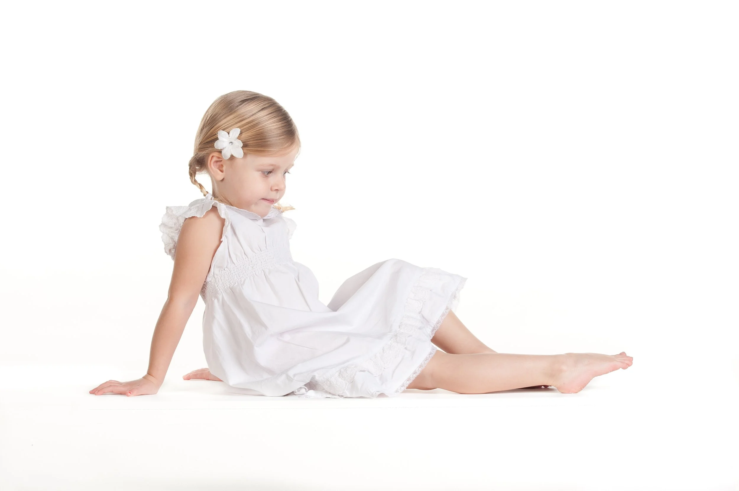 A young girl with blonde hair and a white flower in her hair, wearing a white dress, sitting on the floor against a white background, looking thoughtfully to her left.
