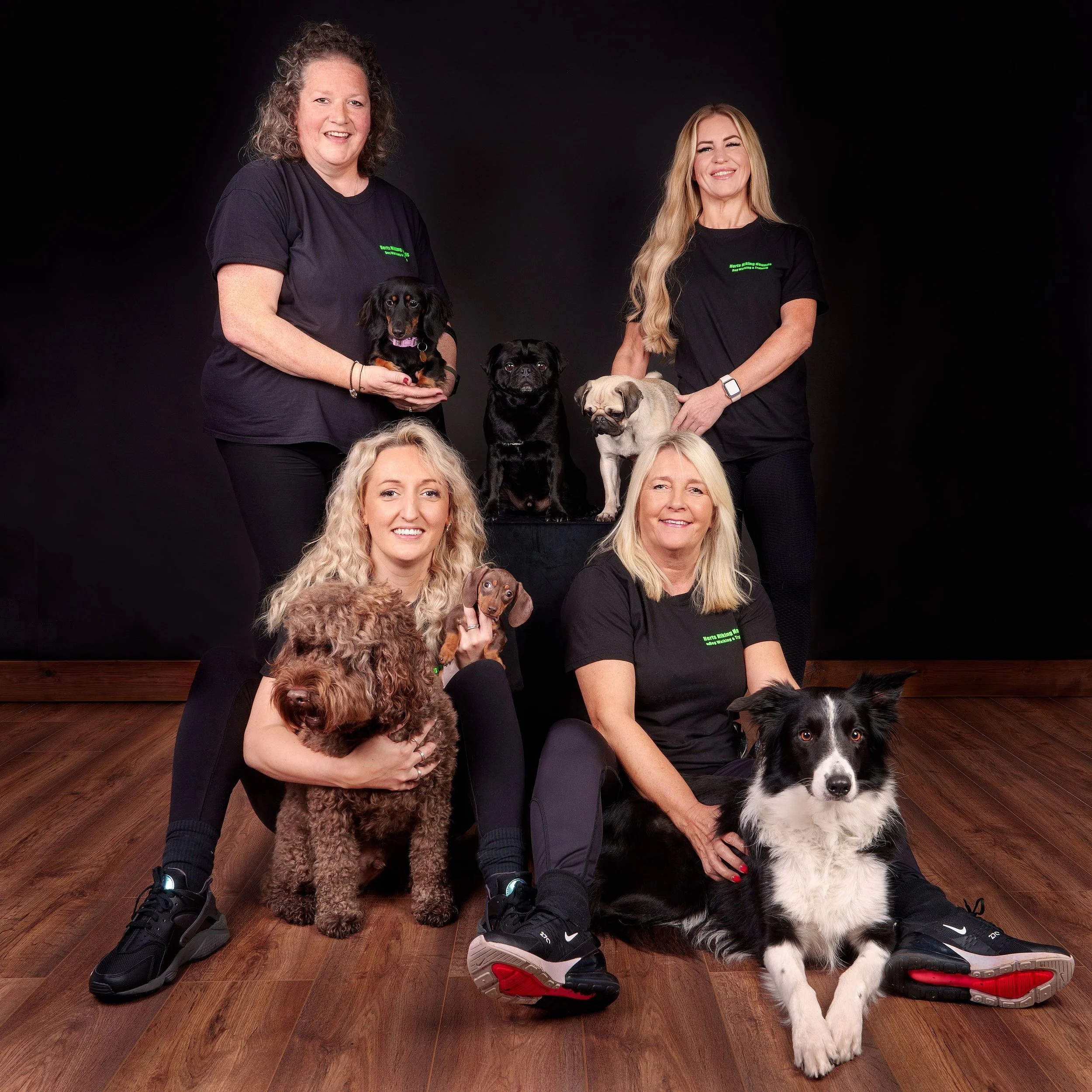 Four women with five dogs posing for a photo against a black background. The women are smiling, and the dogs are of various breeds.