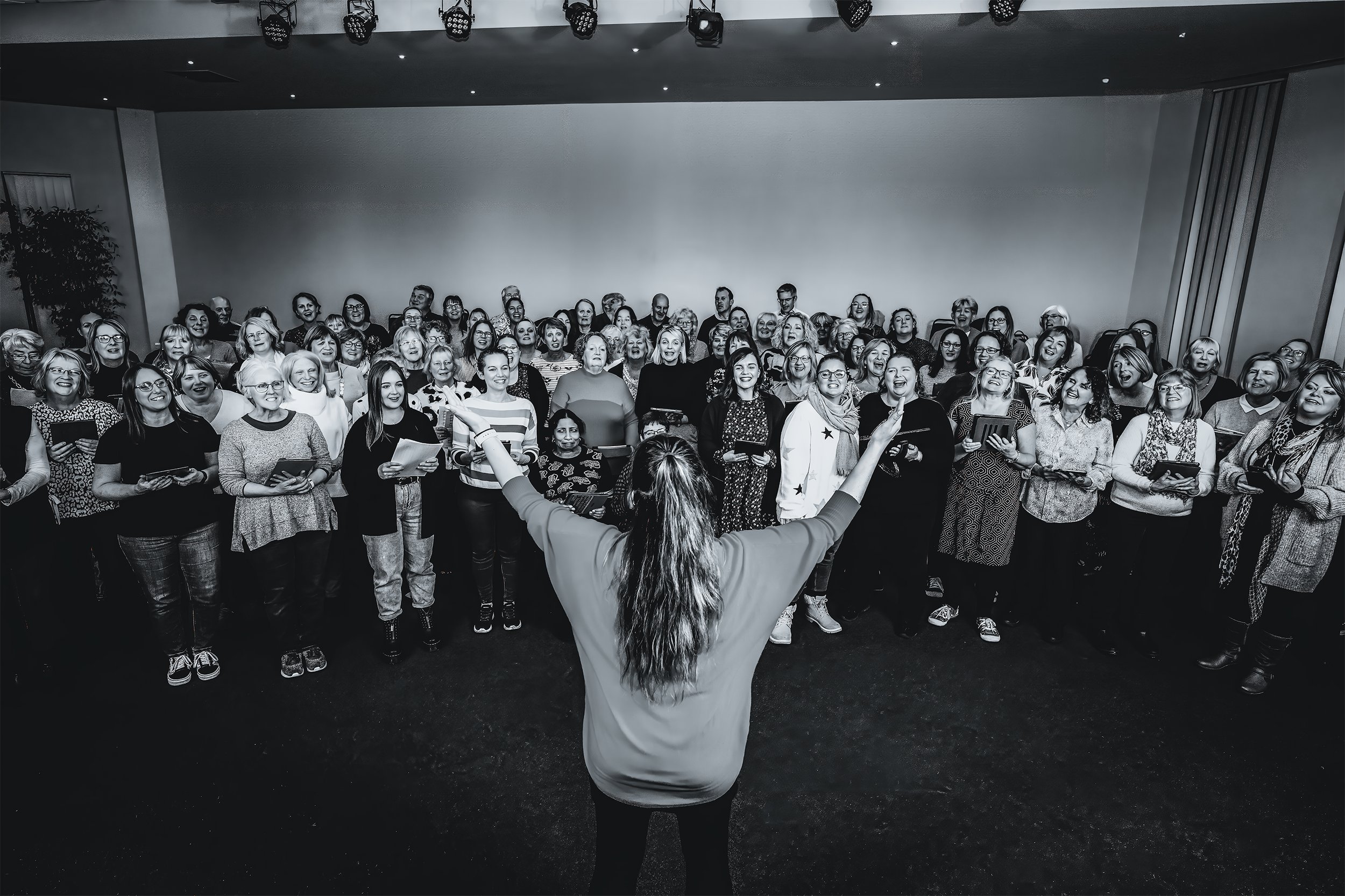 A woman leading a choir or group of people during a performance or rehearsal, with her back to the camera and arms raised, in a large indoor space with stage lighting.