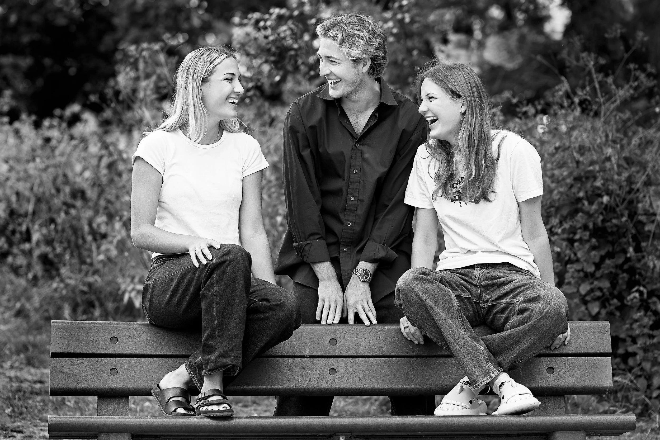 A black and white photo of a man and two young girls sitting on a park bench, all smiling and laughing at each other.