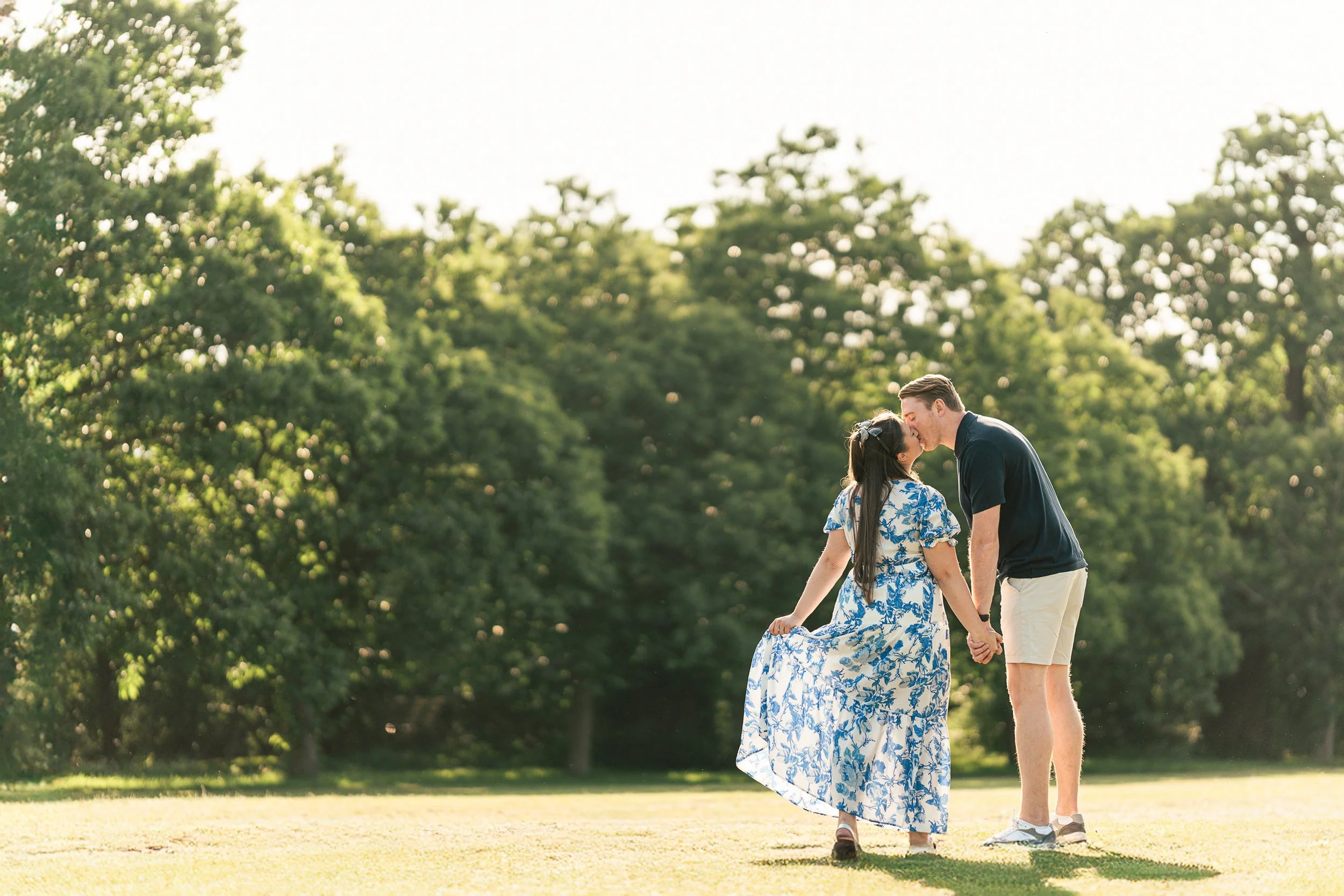 A couple holding hands and kissing in a park with green trees in the background during daytime.