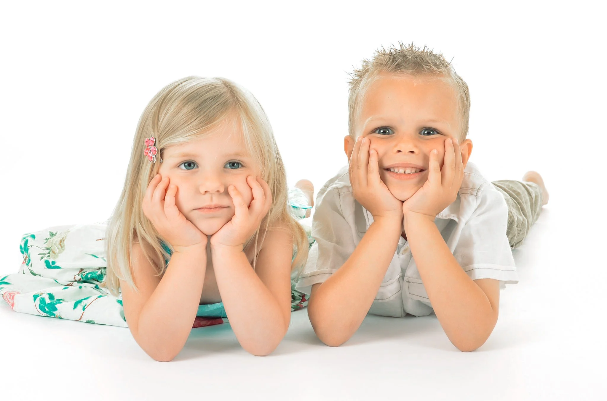 A young boy and girl lying on their stomachs with their chins resting on their hands, smiling and looking at the camera, against a white background.