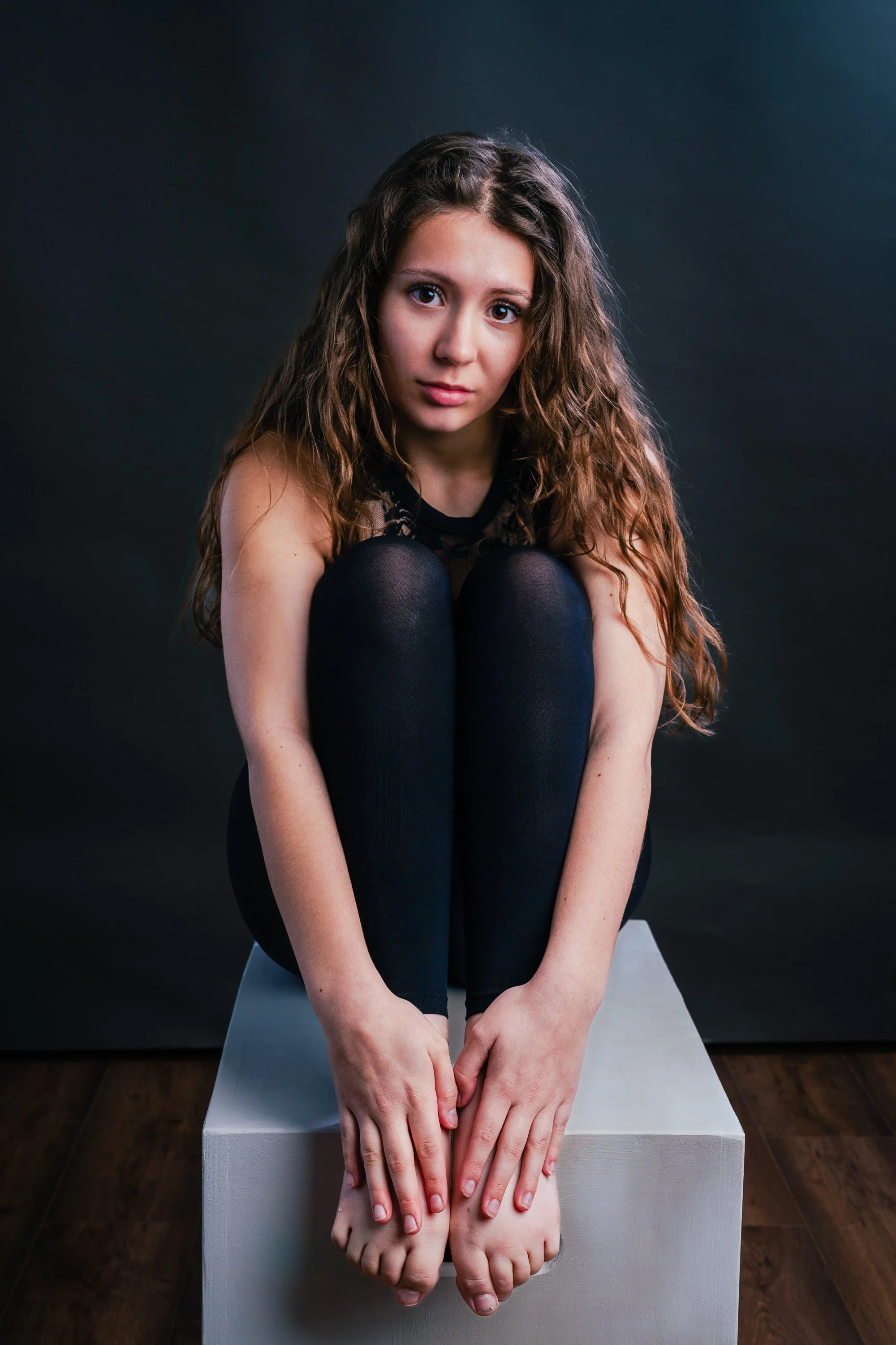 A young woman with long, wavy brown hair sitting on a white platform against a dark background, hugging her knees with her hands, looking directly at the camera.