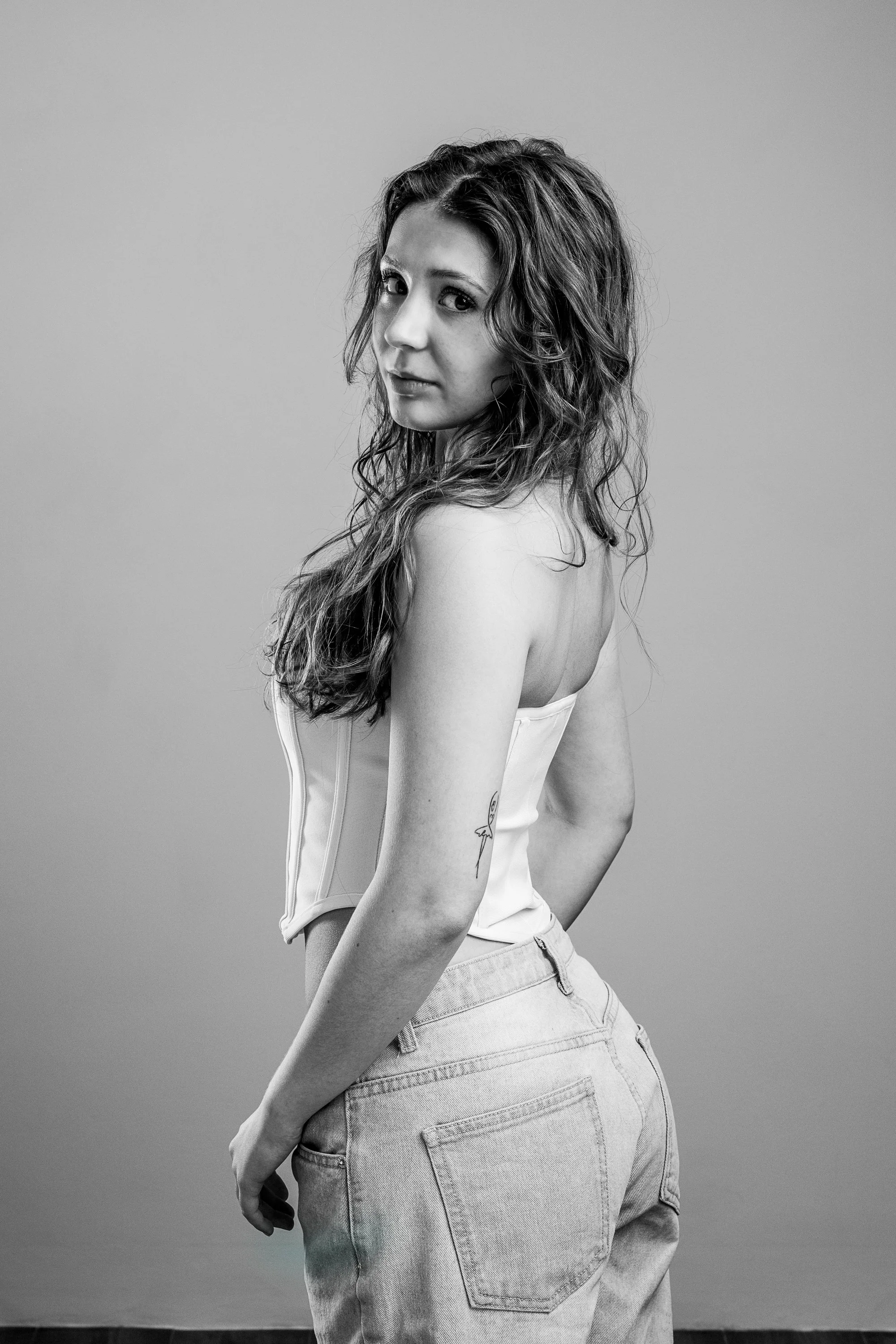 A woman with long curly hair looking over her shoulder at the camera, wearing a strapless top and jeans, black and white photograph.