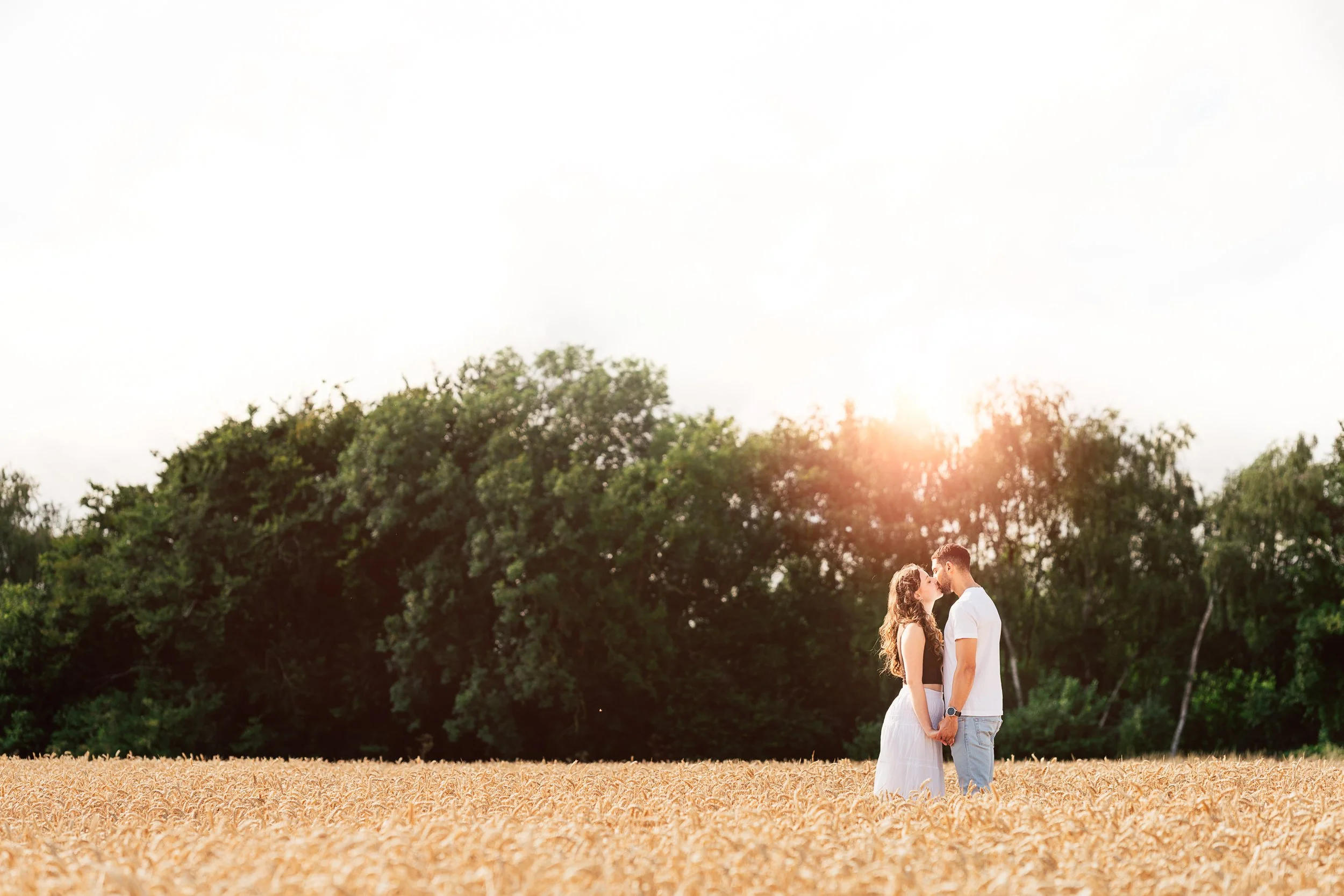 A couple stands in a field holding hands and leaning in for a kiss with a background of trees and a setting sun.