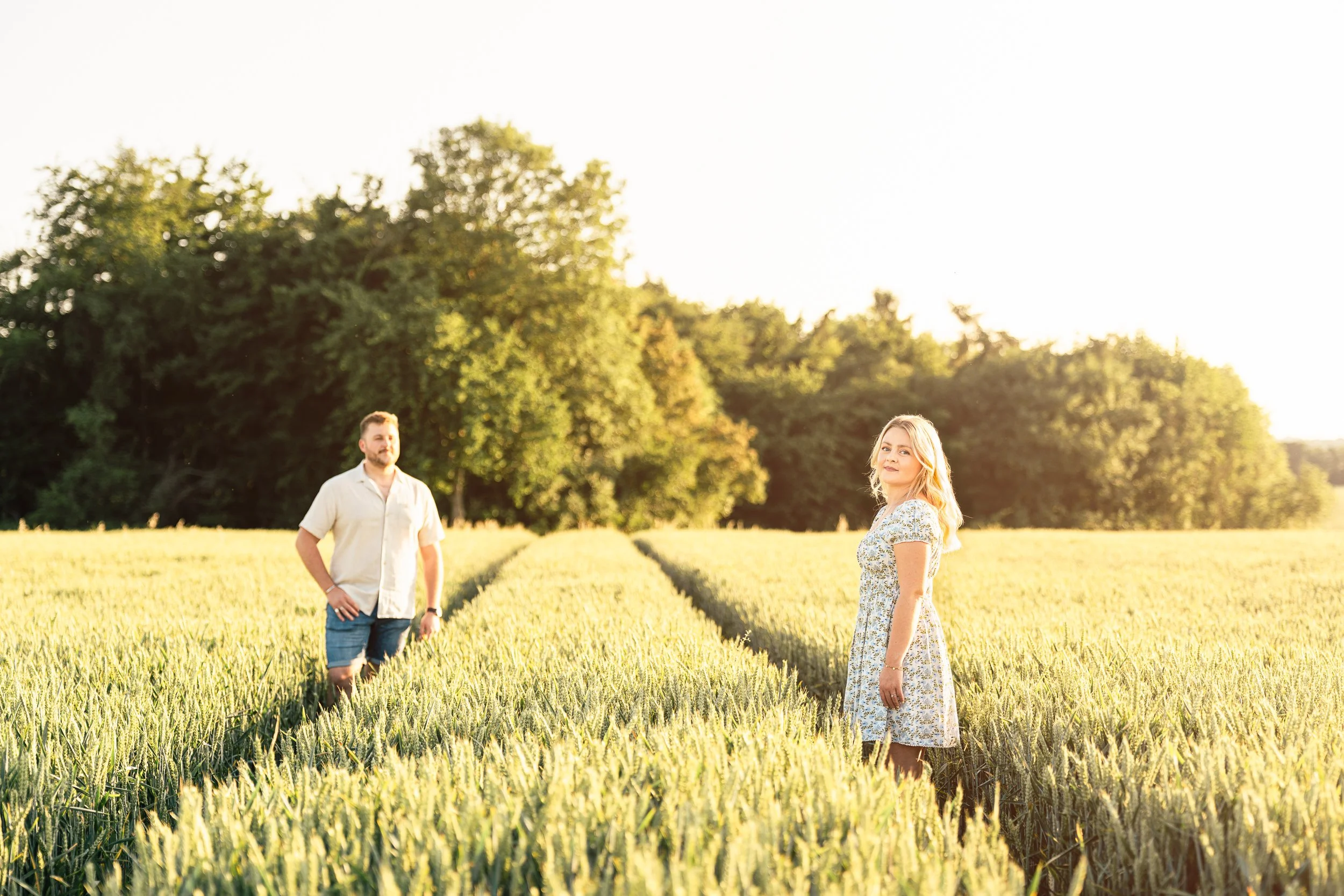 A young woman in a floral dress stands in a wheat field with a man in a light shirt and shorts in the background, both enjoying the golden hour sunlight.