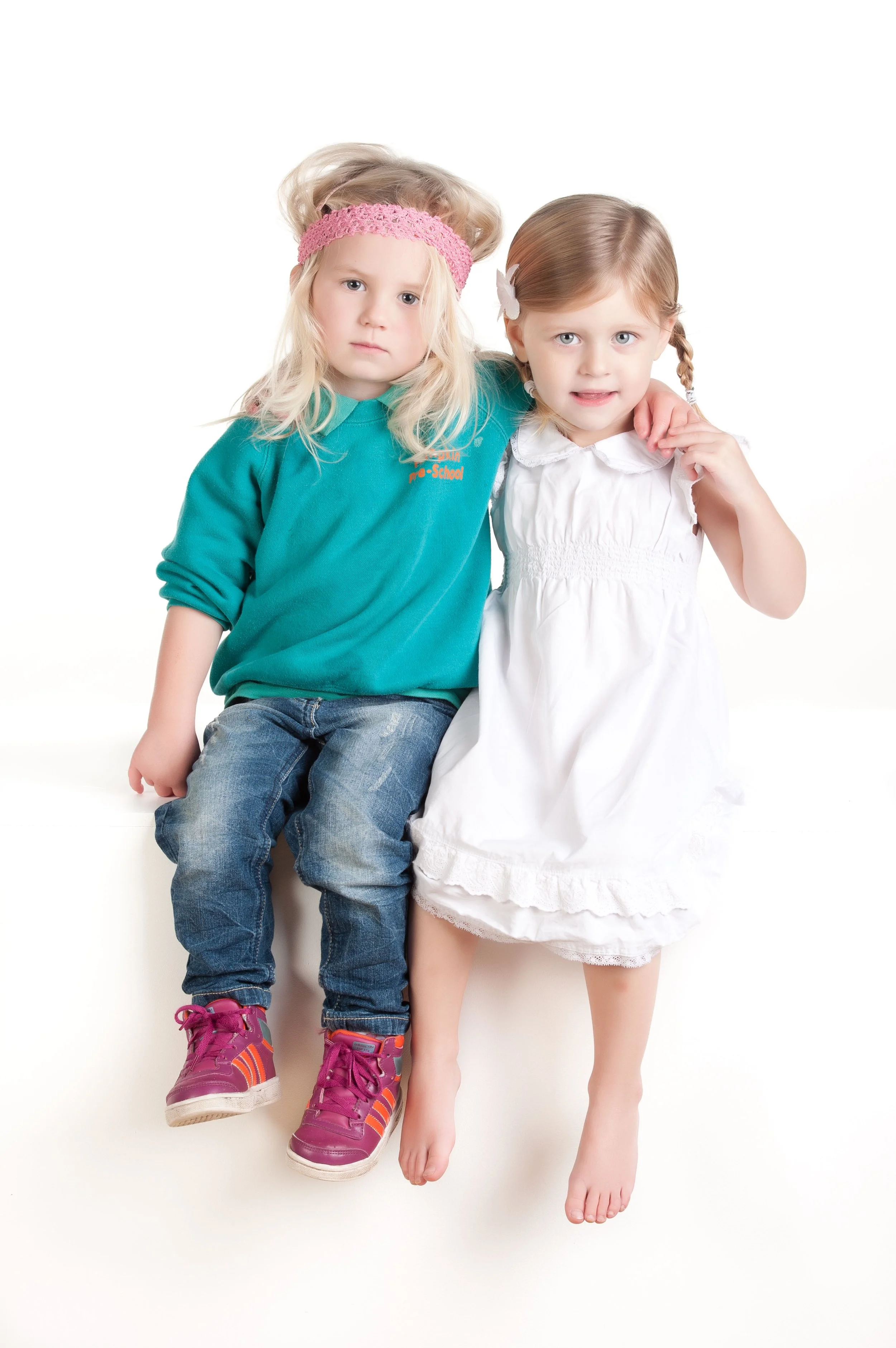 Two young girls sitting together, one with blonde hair and a pink headband, and the other with light brown hair in pigtails, wearing a white dress.