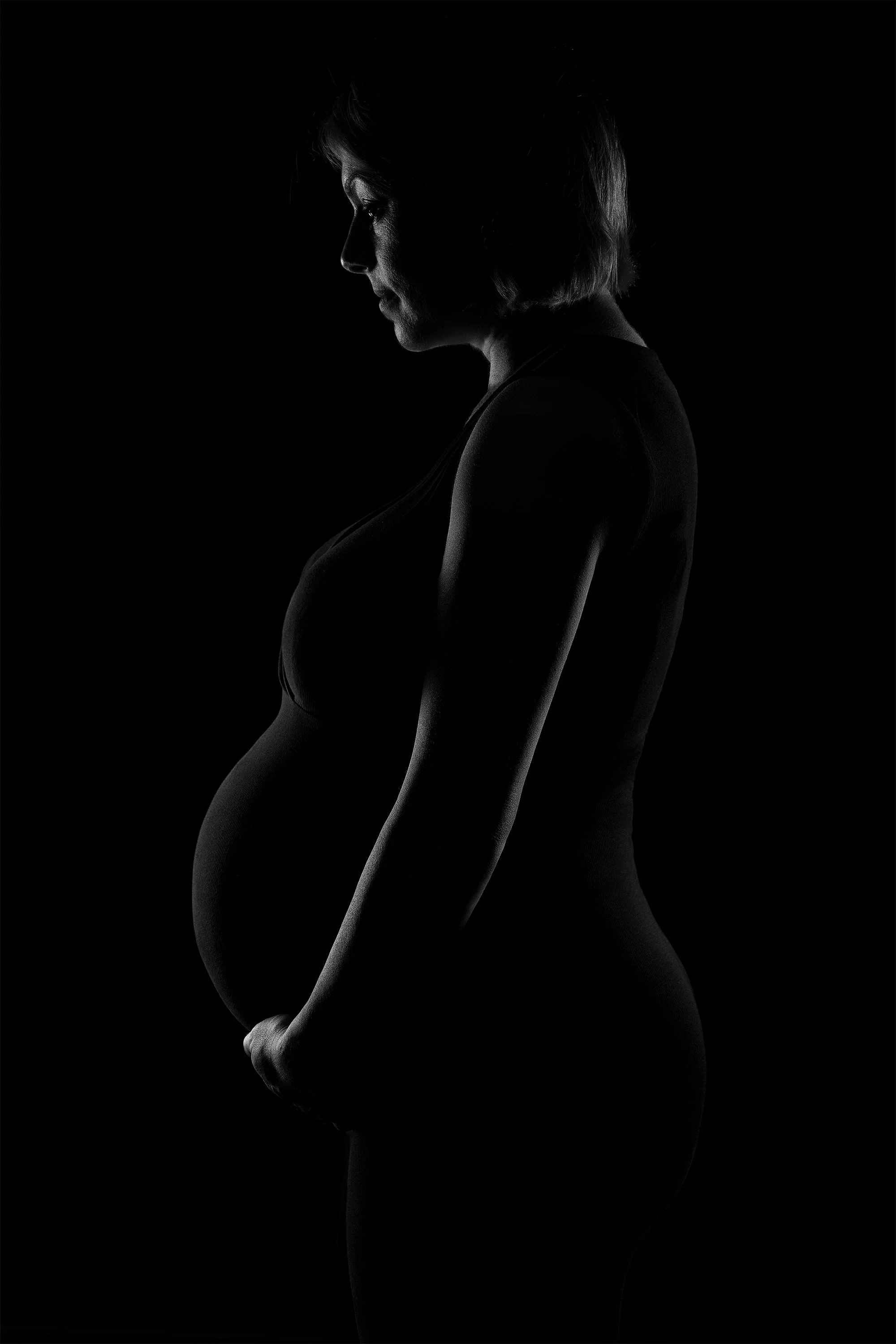 Silhouette of a pregnant woman in profile against a black background, with a soft light highlighting her face, arm, and pregnant belly.