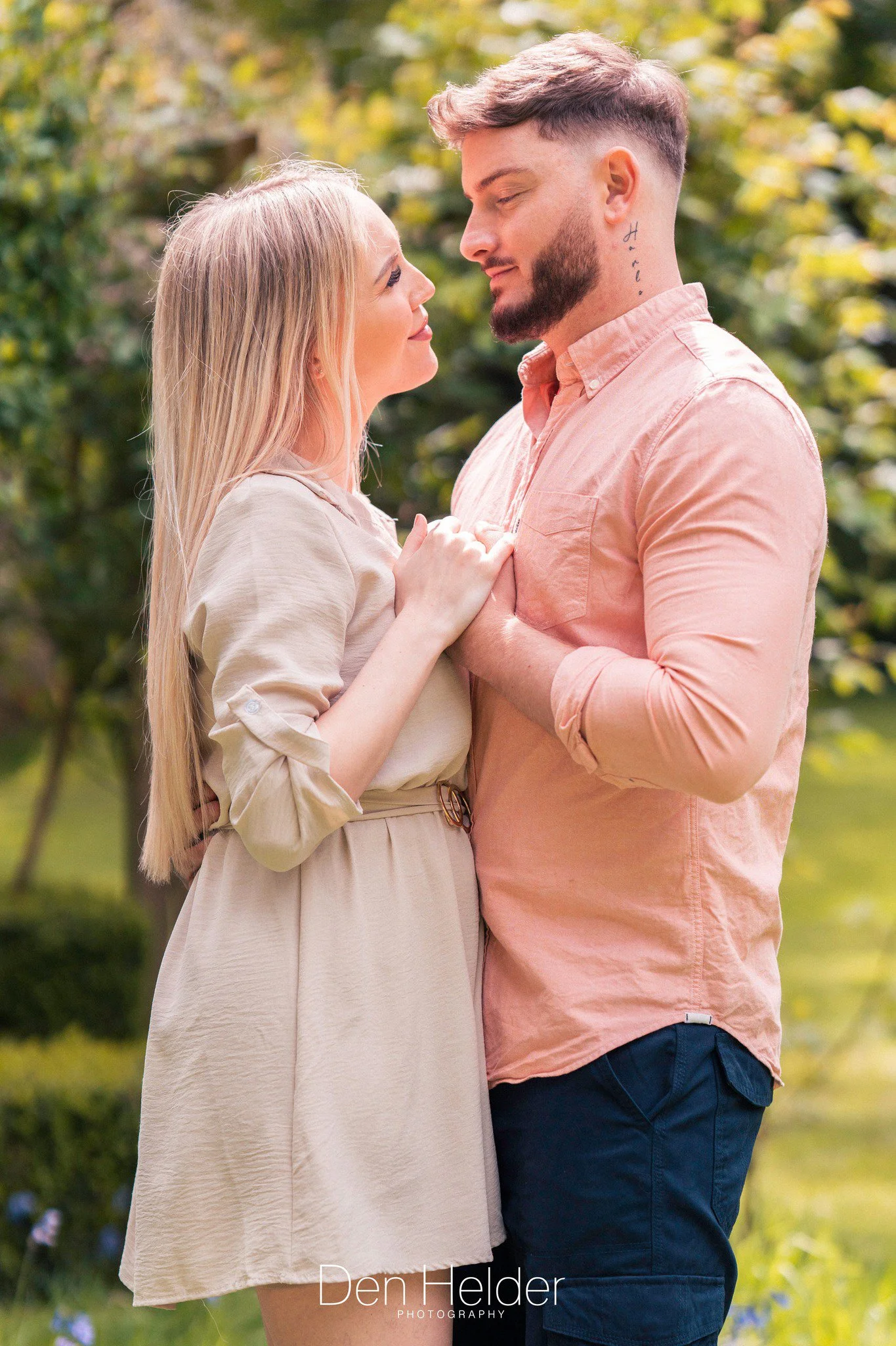 A young couple is standing close together outdoors, gazing into each other's eyes, with their hands gently touching. The background shows green trees and a sunny day.