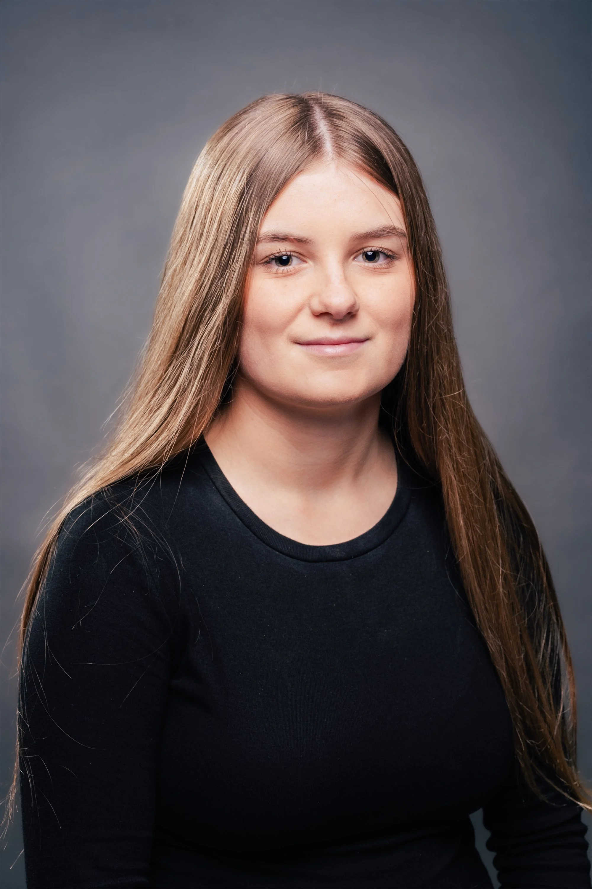Portrait of a young woman with long brown hair, wearing a black top, against a gray background.