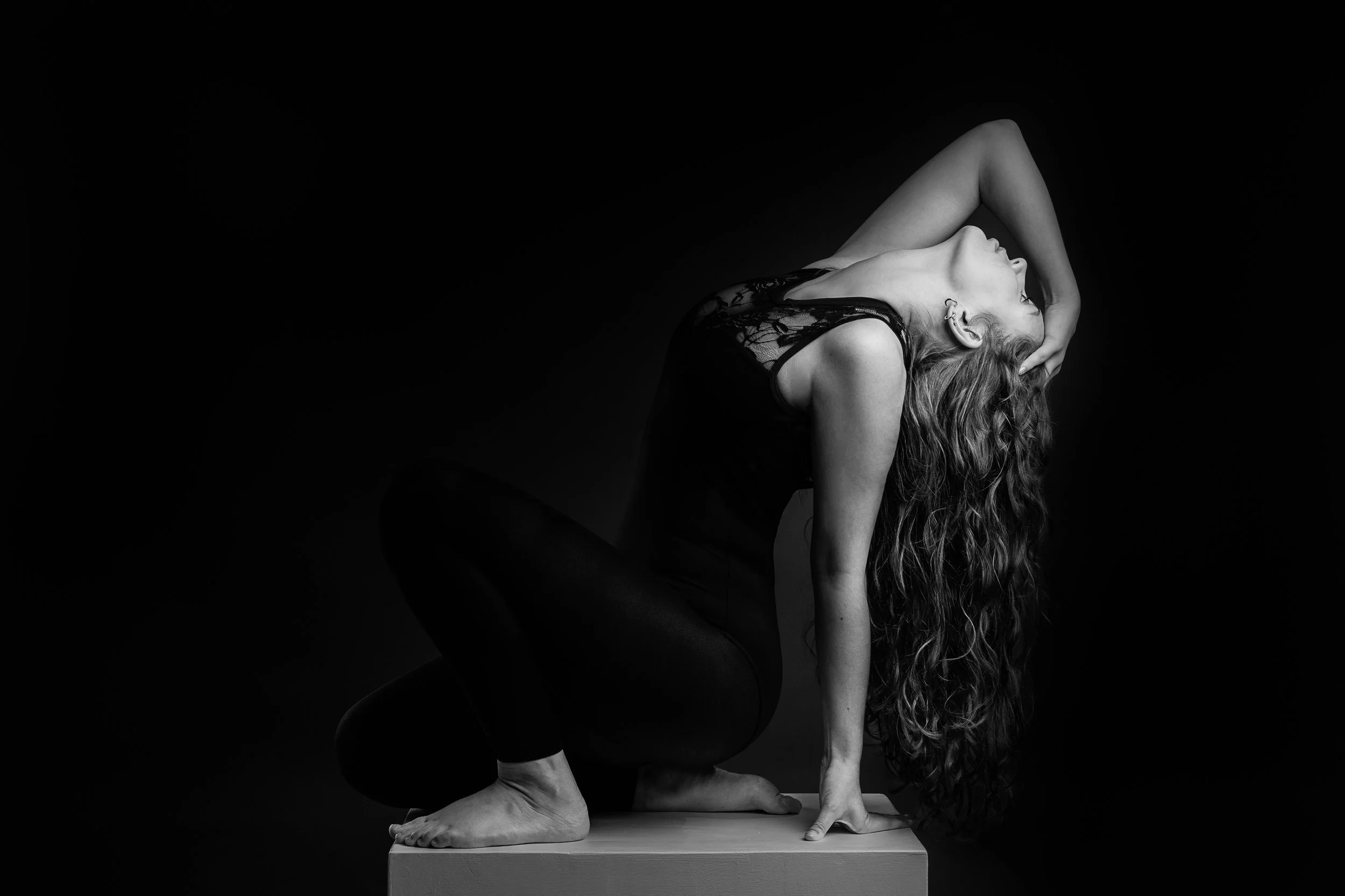 A woman with long curly hair in a black lace top and black pants sits on a box, leaning forward with one hand on the box and the other arm bent over her head, against a dark background.