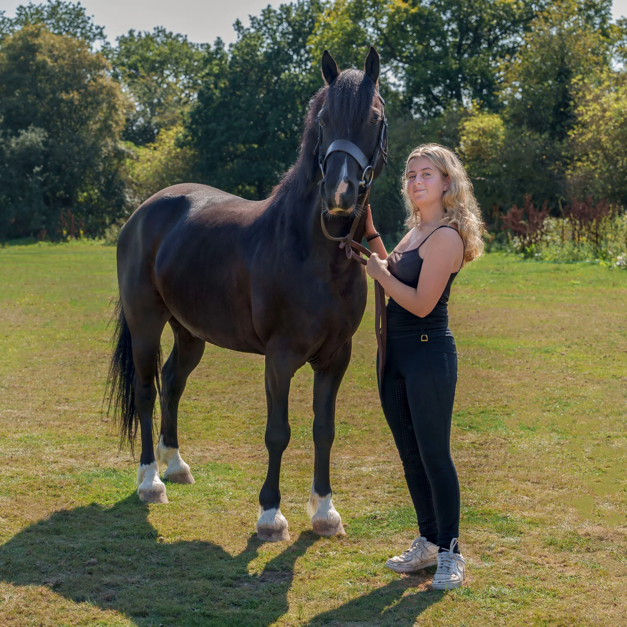 A woman standing outdoors holding the bridle of a black horse with white markings on its legs, with green trees in the background.