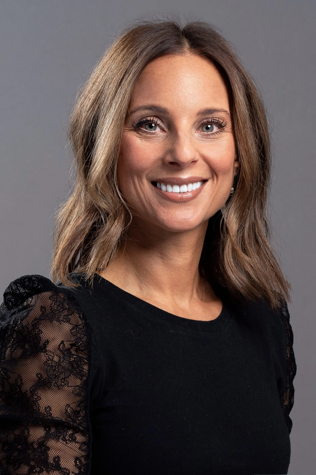 A woman with shoulder-length brown hair, smiling, wearing a black top with lace sleeves, against a gray background.