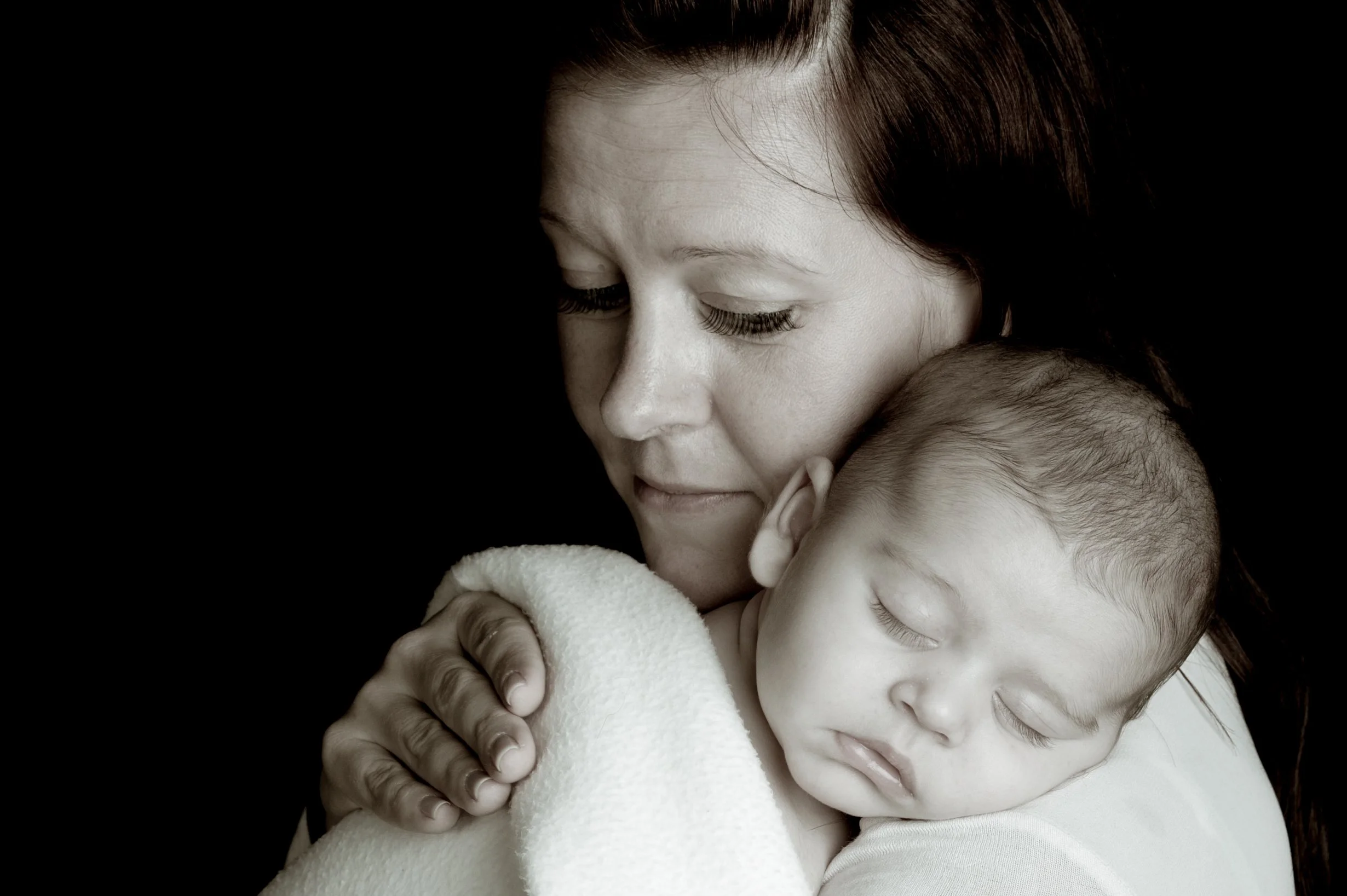 A woman with brown hair holding a sleeping baby with closed eyes, wearing a white blanket, against a black background.